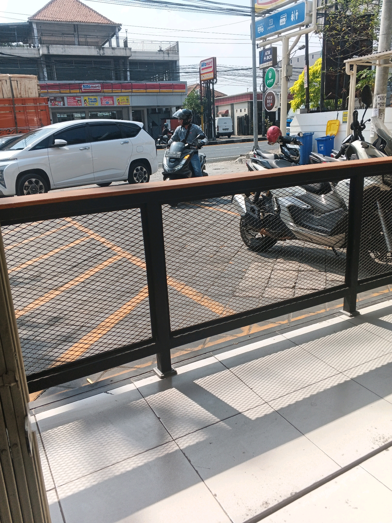 This image depicts a street scene in Genuk Barat, Indonesia. In the foreground, a black metal fence with a wooden top rail runs horizontally across the frame. Behind the fence, a paved area is visible with yellow painted lines, suggestive of parking or traffic markings. Several motorcycles are parked in this area. One person, wearing a helmet and casual clothing, is seated on a black motorcycle. In the mid-ground, a white multi-purpose vehicle is parked, and to its left, a portion of a large orange truck is visible. Dominating the background is a multi-story building with a red tiled roof. Signs for "Alfamart" are prominently displayed on the building's facade, indicating it is a retail store. Additional signage, including a blue sign with icons for "paket", "payment", and "cebit", and another blue sign for "BCA", is also visible on a pole. The environment appears to be a commercial or urban area with clear daylight.