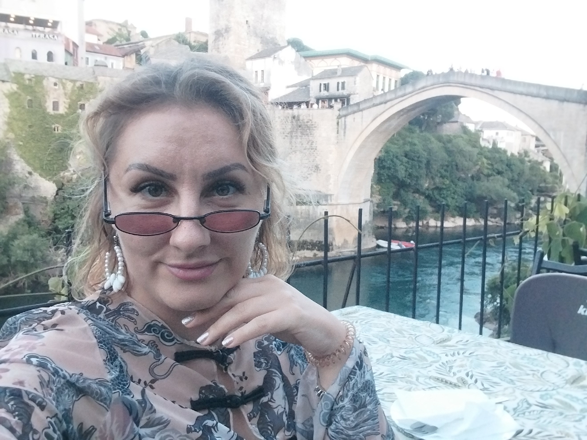 This image captures a woman seated at an outdoor table with the iconic Stari Most (Old Bridge) in Mostar, Bosnia and Herzegovina, visible in the background. The scene suggests a relaxed setting at a riverside cafe or restaurant.
The woman, likely in her 30s or 40s, has medium-length, wavy blonde or light brown hair and is wearing eyeglasses with reddish-tinted lenses. She displays a subtle, closed-mouth smile. Her hand, adorned with sparkly light-colored nail polish and a spiral-shaped bracelet, rests near her chin. She is wearing large white pearl or shell-like drop earrings and a sheer blouse featuring an intricate pattern of dark-colored dragons or mythical creatures on a light, possibly peach-toned, fabric, with a black knotted closure at the collar.
The background prominently features the Stari Most, a historic stone arch bridge spanning the turquoise Neretva River. Several people are visible on the bridge. Flanking the bridge and river are traditional stone buildings with tiled roofs, some partially covered in green ivy, characteristic of Mostar's architecture. A dark metal railing separates the dining area from the river below, where a small red and white boat is visible on the calm water.
In the foreground, a table is covered with a light-colored tablecloth featuring a leafy or floral pattern. A white plate with a napkin sits on the table. Part of a dark grey chair is visible, and the letter "K" from a brand logo can be faintly seen on its backrest. The scene is illuminated by soft, warm light, suggesting late afternoon or early evening, contributing to a serene atmosphere.