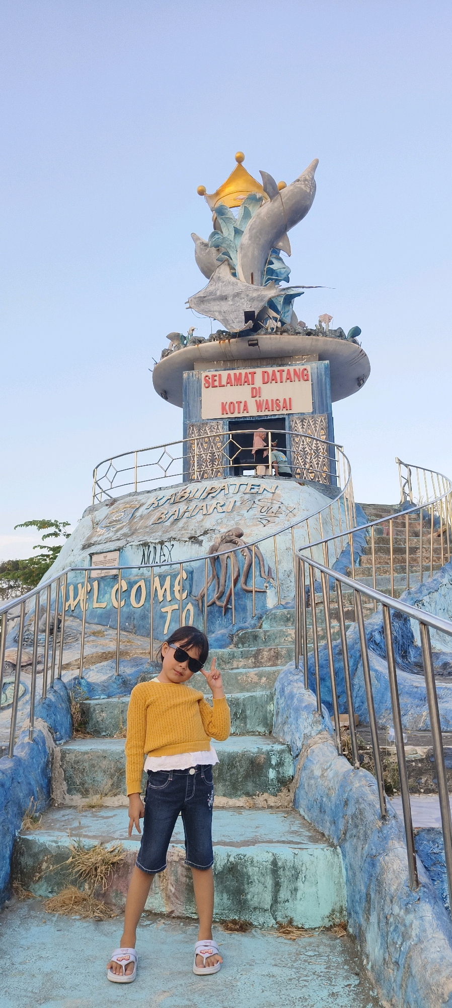 A young girl in a yellow sweater, white undershirt, and denim shorts stands on a blue staircase, striking a peace sign with her right hand and wearing sunglasses. She is positioned in front of a large concrete structure that appears to be a monument or landmark, adorned with blue and white paint and featuring a sculpture of dolphins and a crown at its peak. A sign on the structure reads "SELAMAT DATANG DI KOTA WAISAI," indicating it is an entrance or welcome marker for the city of Waisai, Indonesia. The background shows a clear, pale blue sky. The staircase has metal railings, and some of the concrete structure exhibits wear and tear. A small portion of greenery is visible in the far left background.