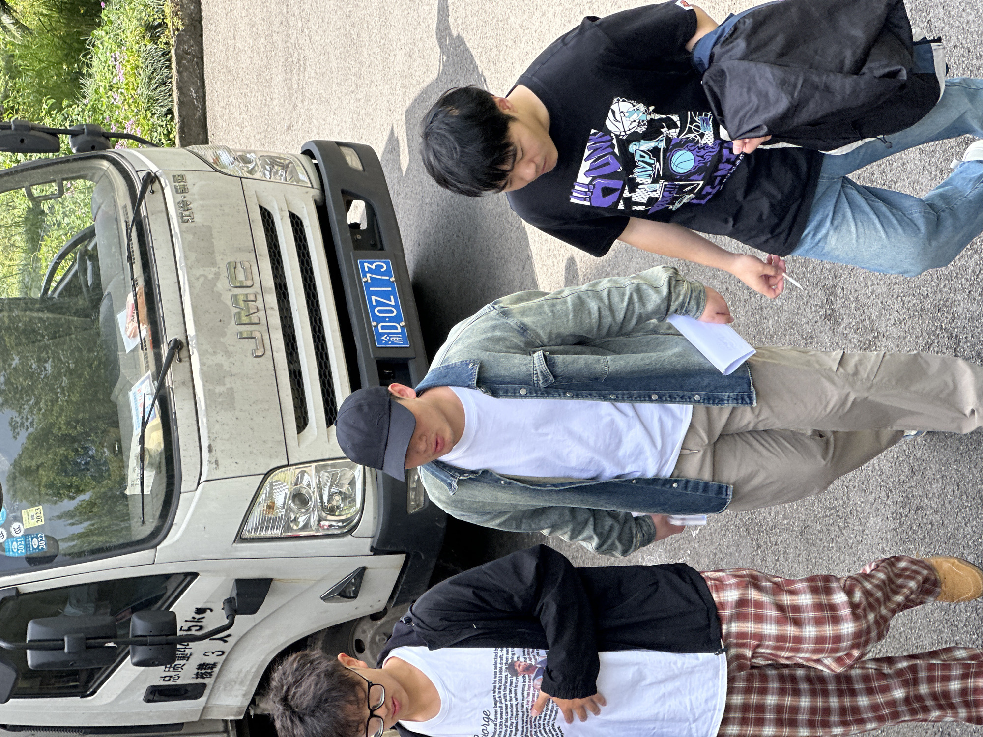 Three young men stand near a white JMC truck in Jiangjin, China. One wears a black shirt with a graphic design, jeans, and carries a black jacket. Another wears a white t-shirt, a denim jacket, khakis, and a cap, holding a piece of paper. The third wears a white t-shirt, a black jacket, and red plaid pants. The truck has a license plate that reads "渝D:OZ173". Text on the truck includes Chinese characters indicating its total mass and passenger capacity. Vegetation is visible in the background.