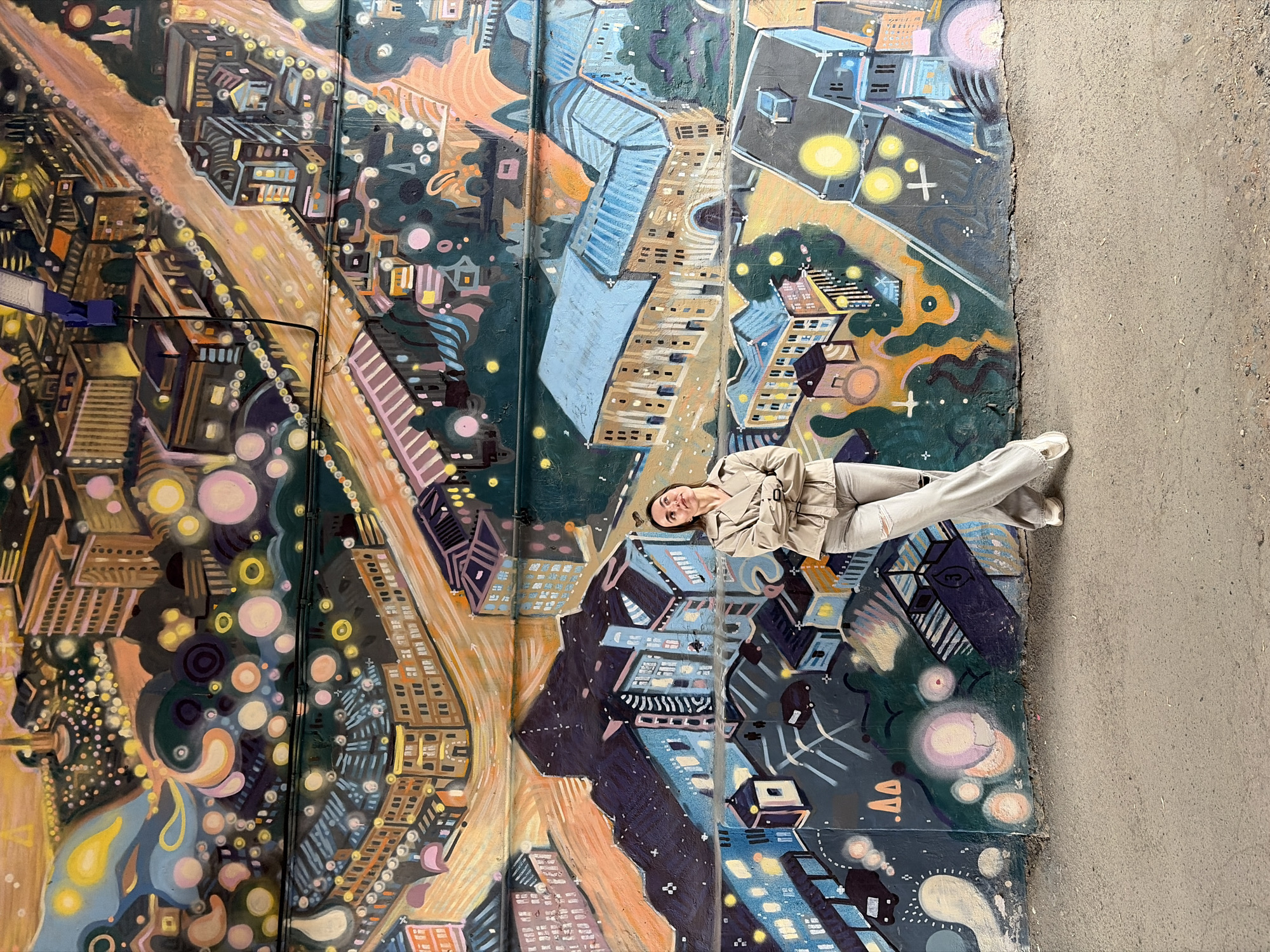A young woman lies diagonally across a painted wall mural in Izhevsk, Russia. The mural depicts a stylized cityscape featuring buildings in a variety of colors including shades of blue, brown, and orange. Streetlights and other elements add to the scene. The woman is wearing a light tan jacket and matching pants. The edge of the mural meets a concrete surface or sidewalk along the right side.