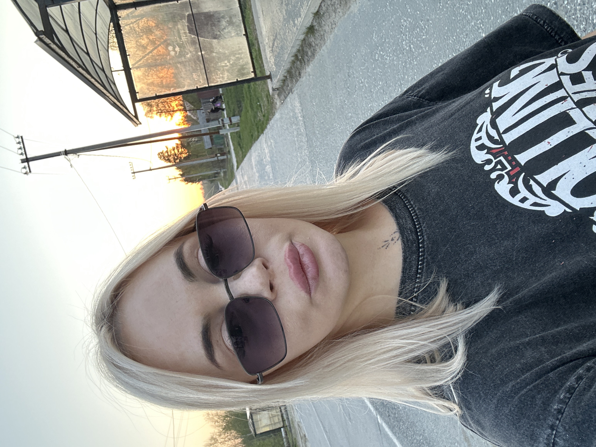 The image shows a woman with blonde hair and sunglasses. She is wearing a dark-colored t-shirt with a white graphic on it. The background suggests an outdoor urban setting, possibly a bus stop or roadside in Высокий, Russia, as indicated by the infrastructure, the presence of a bus shelter, and the presence of utility poles and street lighting. The sun appears to be setting, casting an orange glow.
