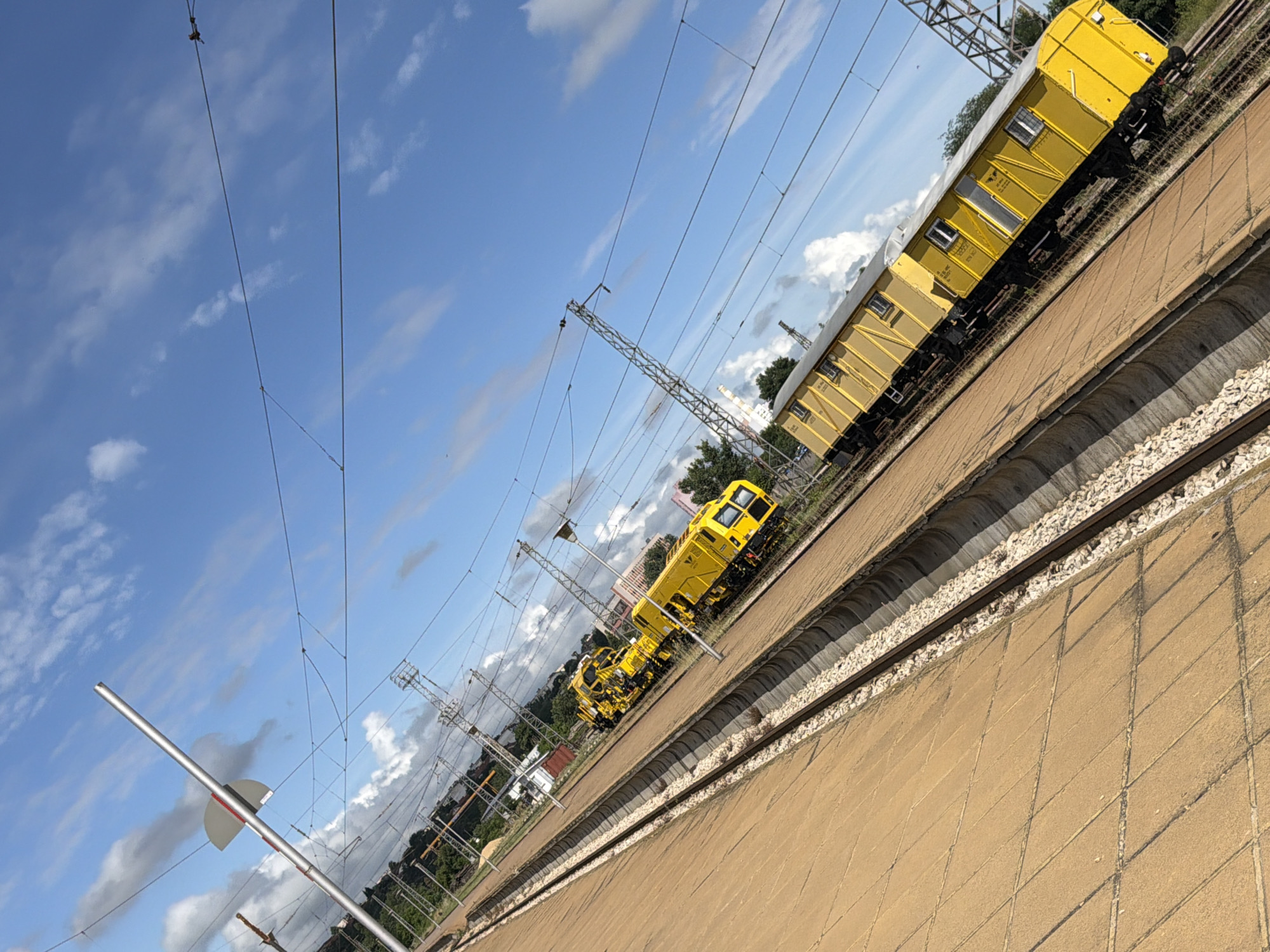 An oblique, eye-level, wide shot captures two yellow railway maintenance vehicles and a yellow railway wagon parked on train tracks under a blue sky with scattered clouds. The scene is likely located in Pleven, Bulgaria, near the railway station, given the infrastructure visible.
In the foreground, the perspective is dominated by the textured surface of a tiled platform, which angles diagonally from the lower left to the upper right of the frame. Adjacent to the platform is a train track, bordered by a concrete curb filled with white ballast.
Further down the tracks, two bright yellow railway maintenance vehicles are positioned side-by-side. The vehicle in the foreground has a more complex structure with visible machinery, while the one behind it appears more like a truck with a cab. Both vehicles are brightly colored, standing out against the muted tones of the tracks and surroundings.
Beyond the maintenance vehicles, a long, yellow railway wagon is parked on another set of tracks. This wagon features several windows and a door on its side, and it also displays some text and symbols.
The background reveals more railway infrastructure, including overhead power lines supported by metal pylons and a signal pole with a red and white flag-like attachment in the upper left quadrant. Beyond the tracks, there are some buildings, trees, and a distant cityscape, suggesting an urban or semi-urban environment. The sky is a vibrant blue, dotted with fluffy white and grey clouds, indicating fair weather. The image is sharply focused, with good lighting that highlights the bright yellow of the vehicles. There are no people visible in the scene.