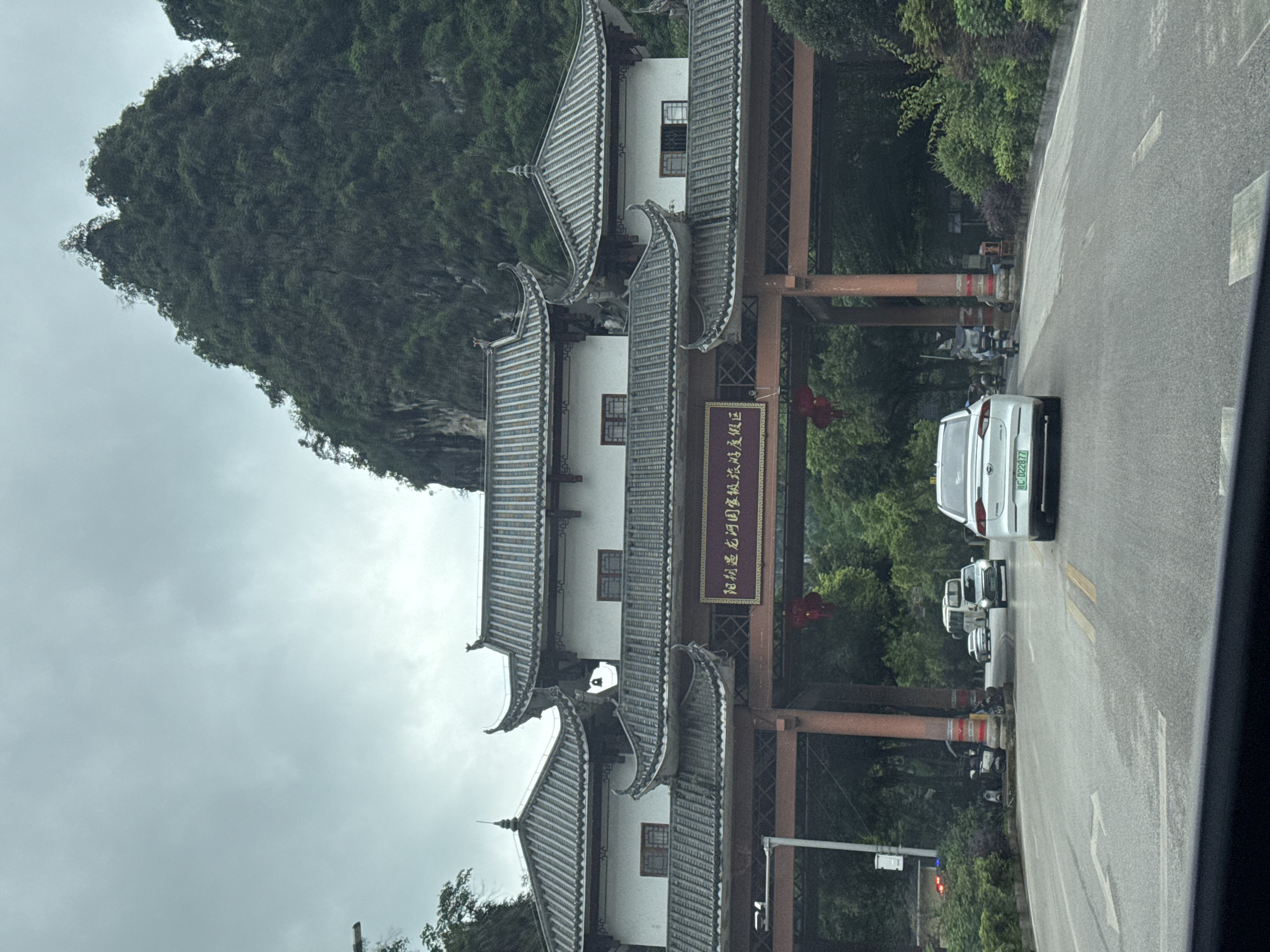 The image shows a traditional Chinese-style gateway structure in Guilin, China. The gateway features multiple tiers of roofs with upturned eaves, typical of traditional Chinese architecture. A sign in Chinese characters is affixed to the structure. The gateway spans a road with vehicles lined up. A forested hill or mountain is visible in the background. The weather appears overcast. A portion of the vehicle's interior that the photograph was taken from is also visible.