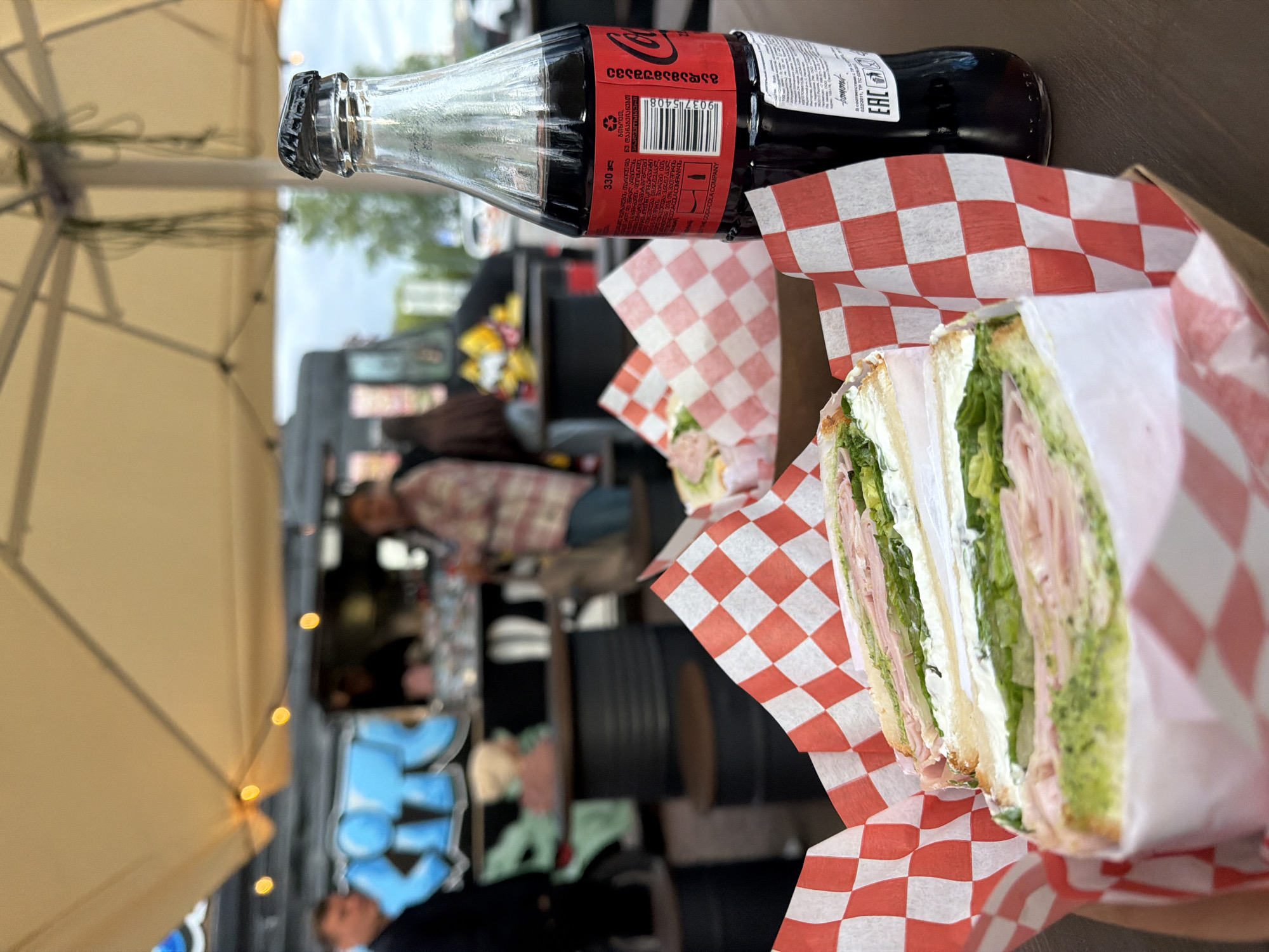 A close-up shot shows a sandwich and a glass bottle of Coca-Cola in a red and white checkered wrapper or box. The sandwich appears to be a stacked deli sandwich with layers of lettuce, cheese, meat, and mayonnaise on white bread. The Coca-Cola bottle is glass and is sealed with a black cap.
The background reveals a blurred outdoor setting, suggesting a food truck area or an outdoor cafe. One or two people are visible in the background, with one wearing a plaid shirt. A food truck or service vehicle is partially visible in the back. A beige-colored umbrella is visible at the top left, and lights are hanging from it.
Based on the text visible on the Coca-Cola bottle, "გადამამუშავე/ECOCACOLACOMPANY," the origin is Georgia.
It is unlikely that this photo was taken in Saint Petersburg, Russia due to this origin and the potential for geopolitical tensions.