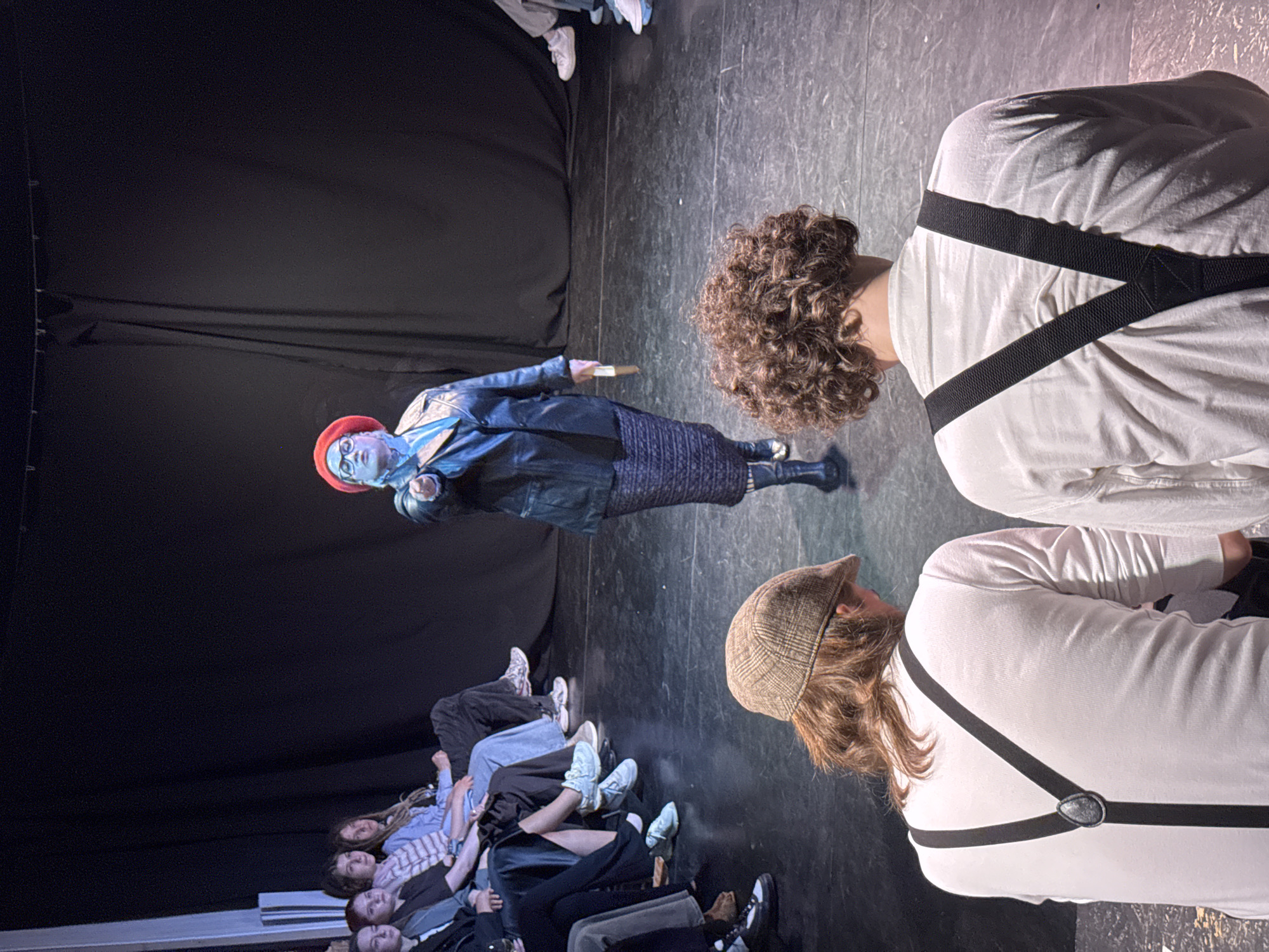 A theatrical performance is taking place. An audience sits on the floor, looking up at a performer wearing a blue painted face, a red hat, a blue jacket, and a skirt. The location is unknown, and there is no indication of the city of Izhevsk, Russia. Two individuals in white shirts with suspenders are seated nearby, facing away from the audience. The stage is dark with black curtains in the background.