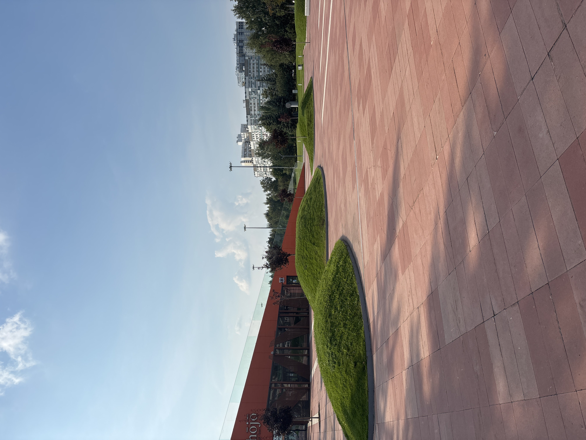 The image depicts a modern public space in Naberezhnye Chelny, Russia, likely a park or plaza, featuring a sleek, orange-colored building with large glass windows and a prominent "hojo" sign. The foreground is dominated by a vast expanse of red brick paving, with dappled sunlight casting shadows across it. Several irregularly shaped, grassy mounds punctuate the paved area, adding a touch of organic design. In the background, a line of mature trees separates the plaza from a cluster of tall, multi-story residential buildings, typical of Soviet-era architecture. The sky is a clear, bright blue with a few scattered white clouds, suggesting a pleasant day. The scene is devoid of people, presenting a quiet and ordered urban environment.