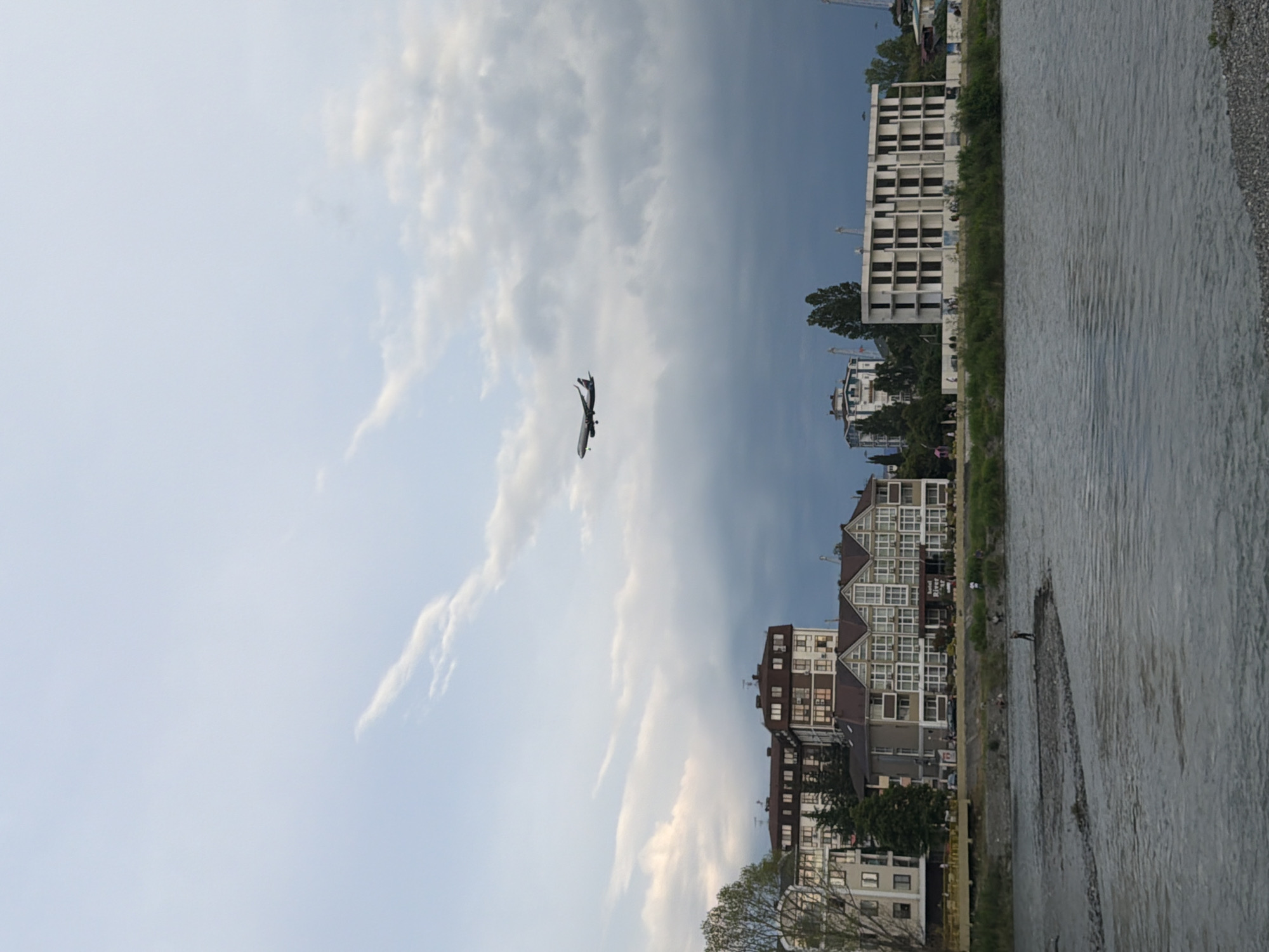 An airplane flies in a cloudy sky above a waterfront city. The city has several multi-story buildings, some with balconies and brown roofs. The buildings are situated along a riverbank, with green grass and trees growing between them and the water. A person is standing in the shallow water of the river. The river is wide and appears to have a rocky bottom. The sky is a mix of light blue and dark gray clouds, with wispy white clouds scattered throughout. The overall scene suggests a coastal city, possibly in a temperate climate.