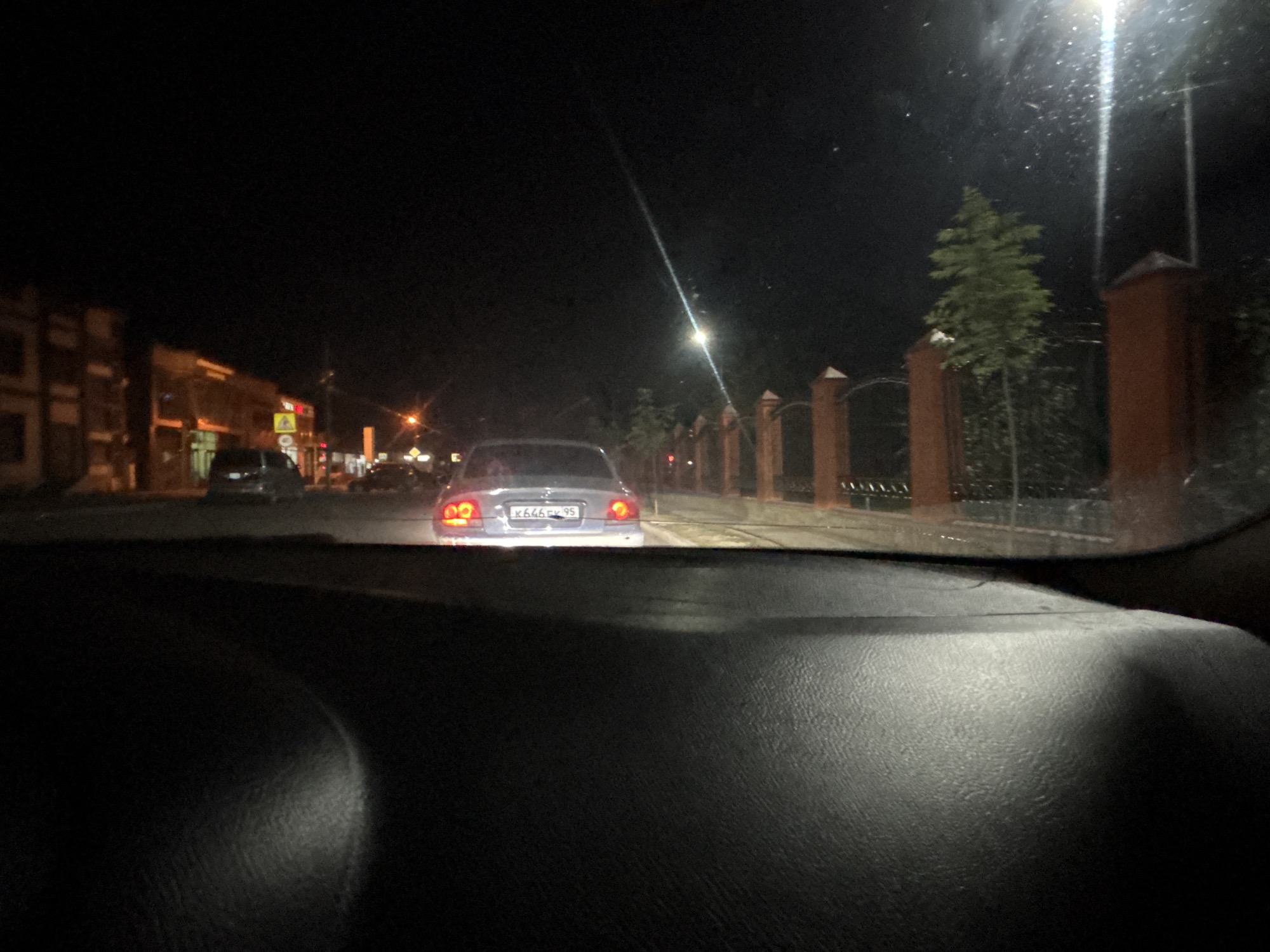 The image shows a nighttime view from inside a vehicle, looking forward through the windshield. The street is illuminated by streetlights and the headlights of other vehicles. In the foreground, the dark, textured dashboard of the vehicle is visible. In the middle ground, a light-colored car with its taillights illuminated is driving away. The license plate of this car is partially visible, appearing to be "K646FK 95". To the left, buildings with illuminated windows line the street, suggesting a commercial or residential area. To the right, a fence with brick posts and a small tree are visible, along with a bright streetlight that creates a lens flare effect. The overall environment appears to be a city street at night, possibly in Grozny, Russia, given the license plate format.