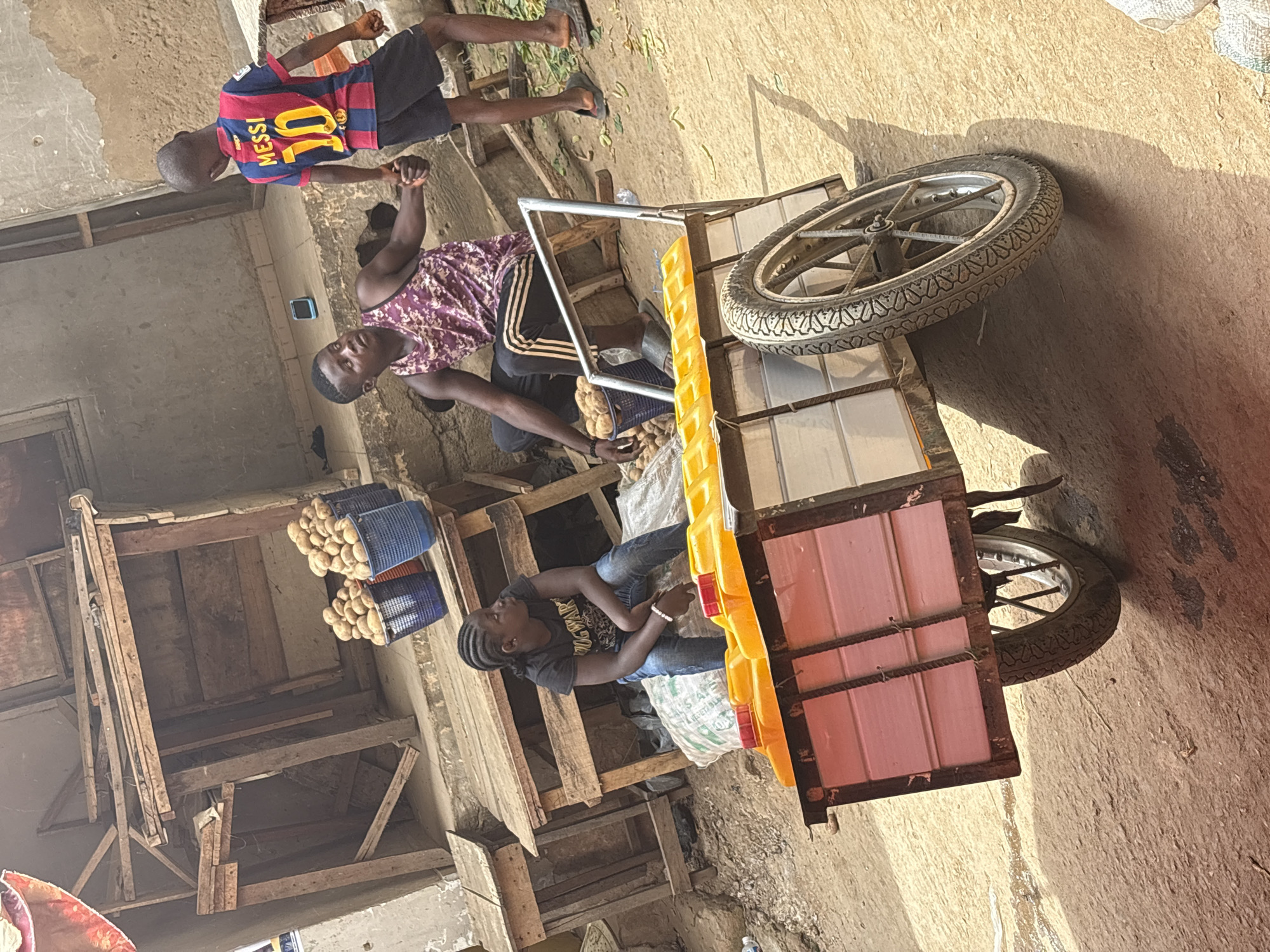 This image captures a vibrant street or market scene in Iddo Tudunwada, Nigeria, under bright, sunny conditions, indicated by strong shadows. The setting features a dusty or unpaved ground and makeshift wooden structures that appear to be market stalls or storage areas.
There are three main individuals in the frame. In the upper left, a young boy, possibly between 6 and 10 years old, stands wearing a blue and red striped football jersey with "MESSI" and the number "10" clearly visible on the back. He wears dark shorts and sandals. Near the center, a man with a patterned sleeveless shirt stands next to a three-wheeled cargo cart, which forms a prominent object in the midground. He appears to be looking towards the left, perhaps at the wooden structures or the items on display. Another man, with braided hair, sits on a large white bag on the cart, wearing a black t-shirt and blue jeans, also looking towards the left. All individuals have neutral expressions.
The cart features a metal frame, a large spoked wheel with a patterned tire in the foreground, and pink paneling at the rear with a yellow top or tray. Several blue plastic baskets filled with light-colored produce, likely potatoes, are visible on the wooden structures and possibly on the cart itself, suggesting a vendor's setup. A large white sack, possibly containing more produce, is used as a seat by the sitting man, and some blurry text including "VEGETAB" can be discerned on it. The overall activity appears to be related to commerce, with the individuals present in what seems to be a daily market environment.