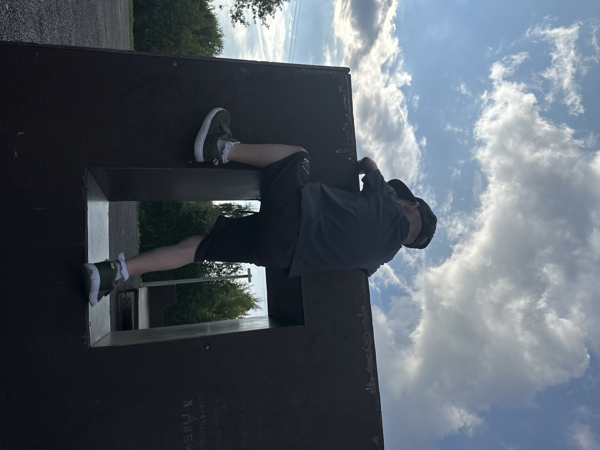 A young boy wearing a black baseball cap, dark t-shirt, and shorts is climbing over a dark, rectangular structure outdoors. His right foot is placed on the top edge of the structure, and his left leg is bent, with his left foot near the bottom of a rectangular opening in the structure. He is facing away from the camera, towards a sky filled with white and grey clouds. The background shows trees and a paved area. The structure appears to be part of an outdoor obstacle course or playground. The scene is captured from a low angle, looking up at the boy and the sky. The location is identified as Chaikovsky, Russia.