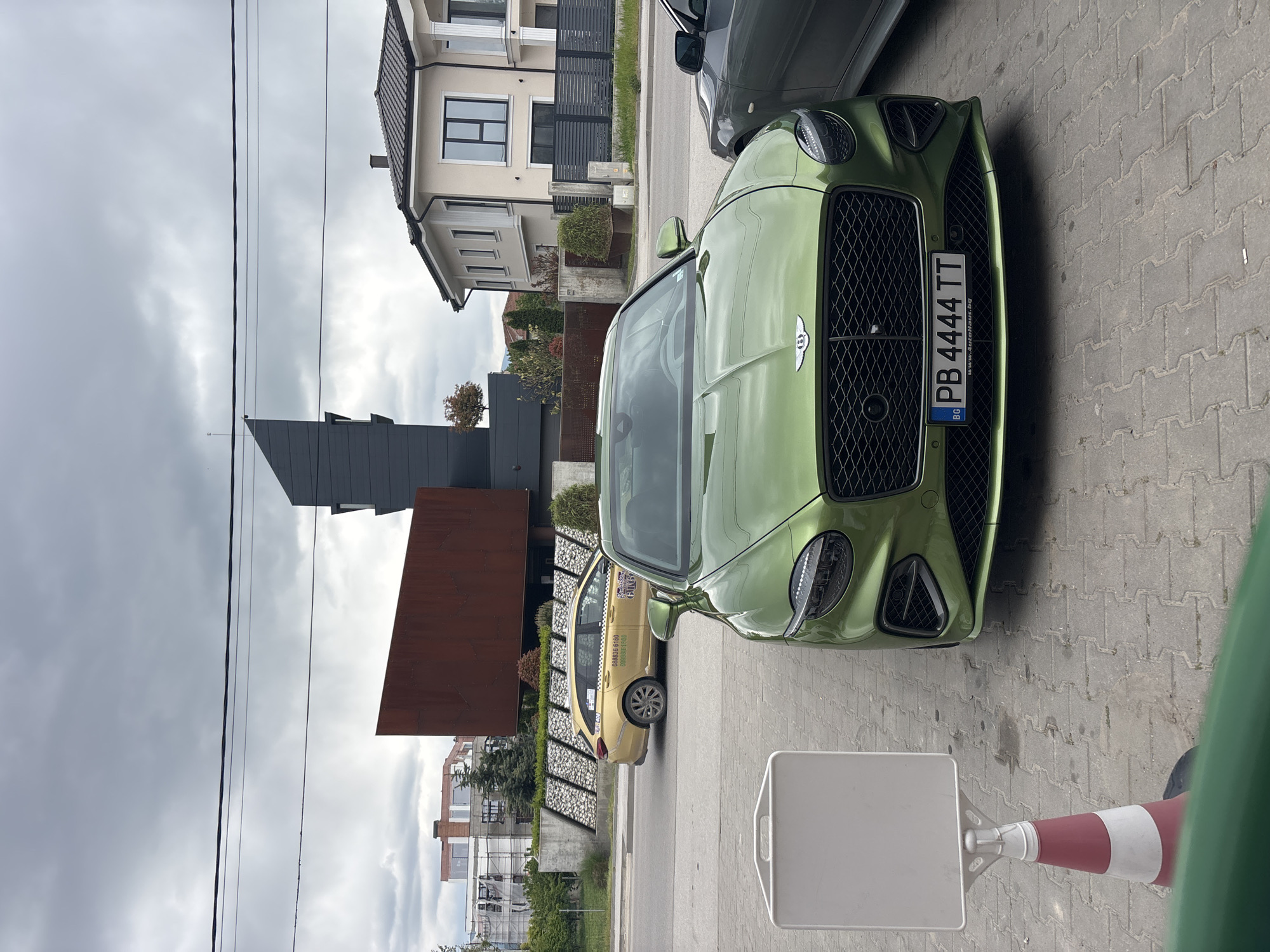 A green Bentley Continental parked on a brick pavement in Plovdiv, Bulgaria, is the scene's focus. The license plate reads "PB 4444 TT." A yellow taxi is parked behind the Bentley. Modern architecture is visible in the background, including a building with a dark, angular design and a structure with rusty brown panels. Overhead power lines run across the cloudy sky. A white sign and a traffic cone are in the foreground. Another car is partially visible on the right, parked next to the Bentley.