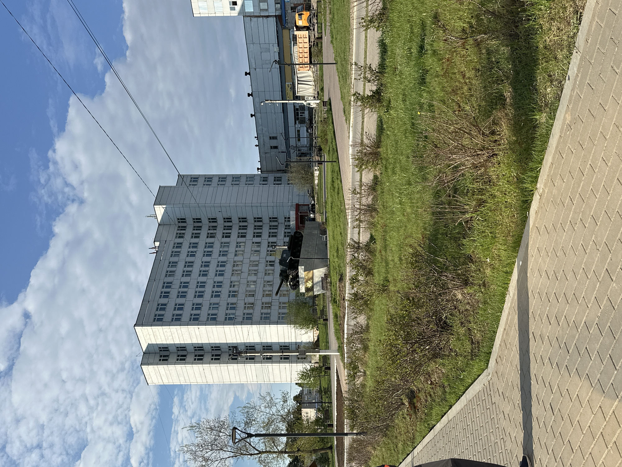 The image depicts a view in Chaikovsky, Russia. In the foreground, there is a paved walkway made of rectangular blocks. Next to the walkway is a grassy area with bushes and some trees. Beyond that stands a tall, white, Soviet-era building with numerous windows, likely a residential or administrative structure. A military memorial featuring a tank is in front of the building. The sky above is blue with scattered white clouds. In the background, there are additional buildings and some vehicles.