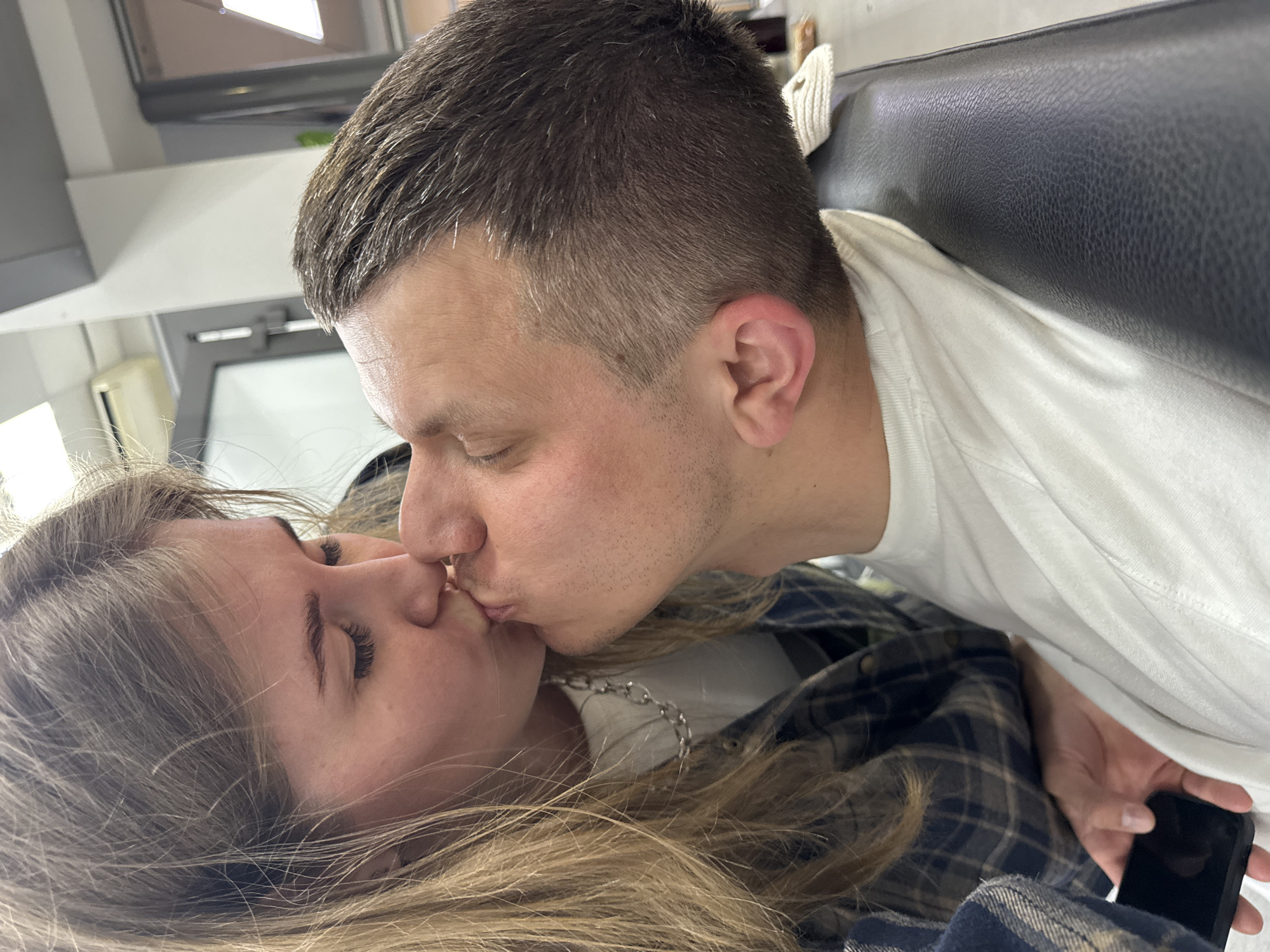 A photograph depicts a man and a woman kissing indoors. The woman has long, blond hair and is wearing a plaid shirt. The man has short, dark hair and is wearing a white t-shirt. The location appears to be inside a vehicle, with a black leather-like seat visible. The image might be related to the city of Perm, Russia, as per the instructions. The man is holding a phone in his right hand.