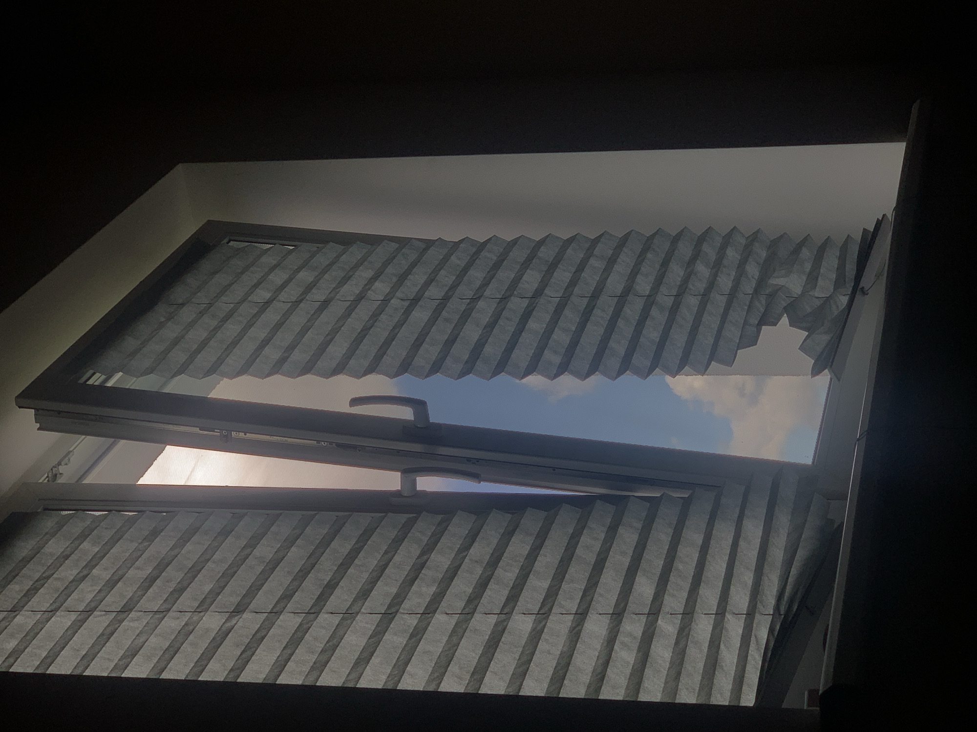 An interior shot captures a partially open window with pleated blinds. The left blind is mostly closed, while the right blind is open, revealing a bright sky with clouds and the interior of a room with dark walls. A window handle is visible. The scene appears to be a residential interior, likely in Minsk, Belarus, given the context of previous interactions.