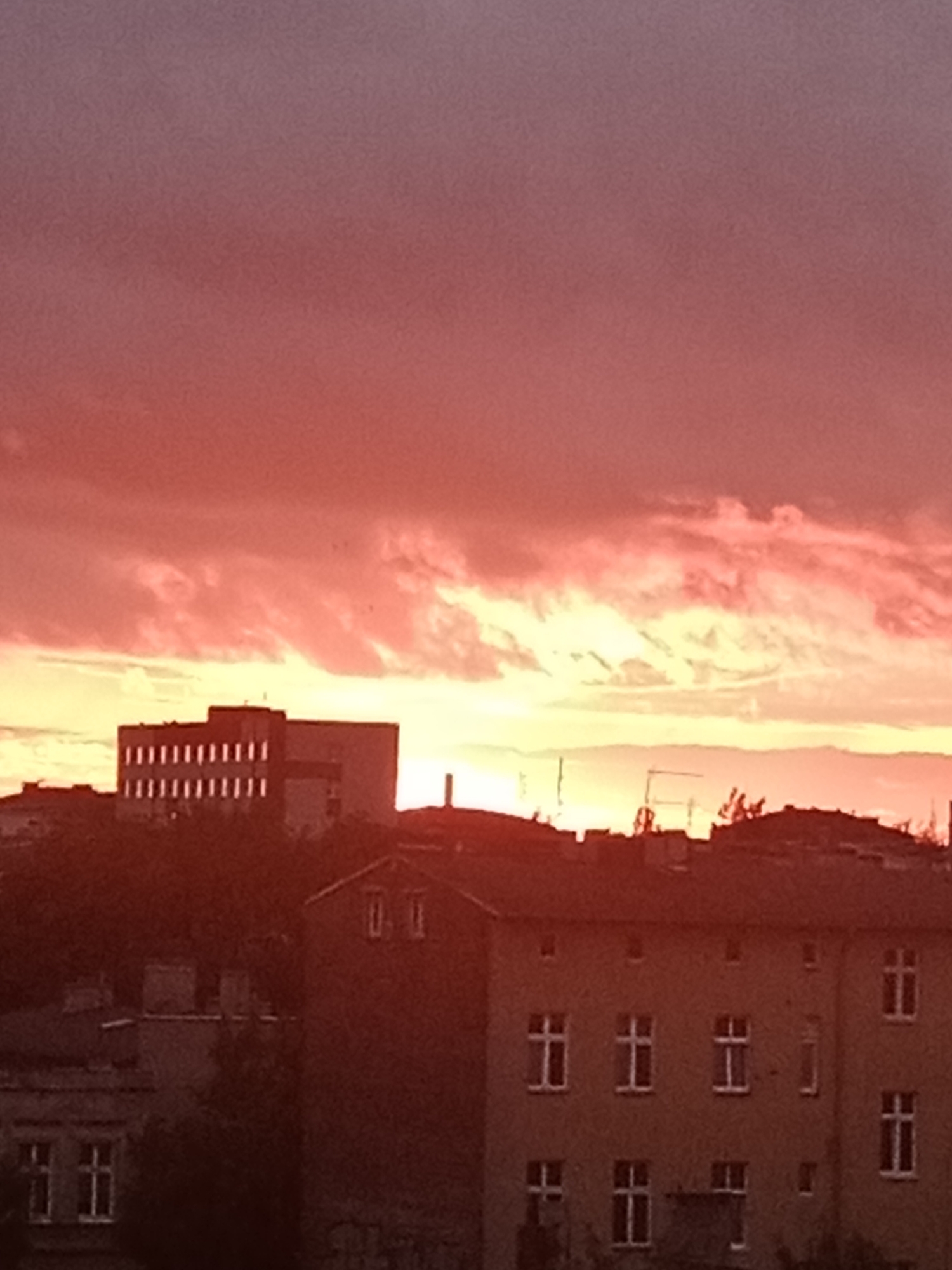 The image captures a vibrant sunset over Bydgoszcz, Poland. The sky is dominated by hues of deep red, orange, and yellow, with clouds streaking across the horizon. In the foreground, several buildings are silhouetted against the colorful sky. The structures appear to be apartment buildings, with visible windows and rooflines. The architecture suggests an urban setting. The overall impression is one of a peaceful evening in the city, illuminated by the dramatic colors of the setting sun.
