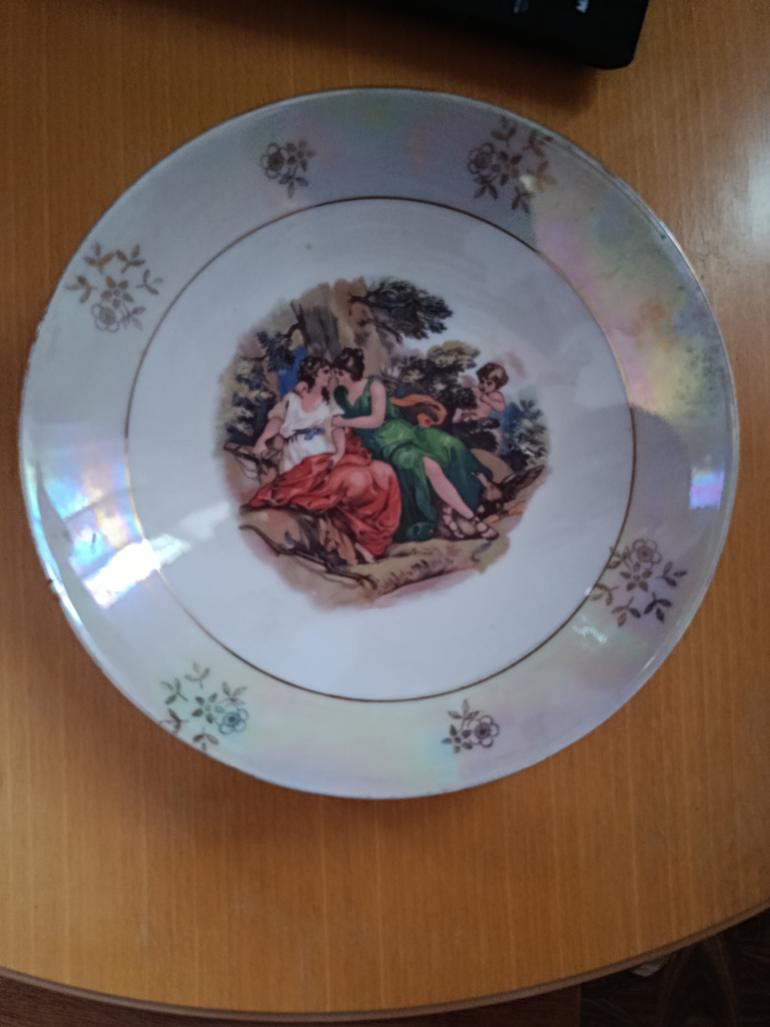 A decorative plate sits on a wooden surface. The plate features a central image of three figures in classical attire, possibly women and a cherub, in a natural setting with trees and rocks. The rim of the plate is decorated with a pearlescent glaze and a gold band, with small floral motifs repeated around the edge. The wooden surface appears to be a tabletop or desk. The image does not provide any information about the city of Voznesensk, Ukraine.