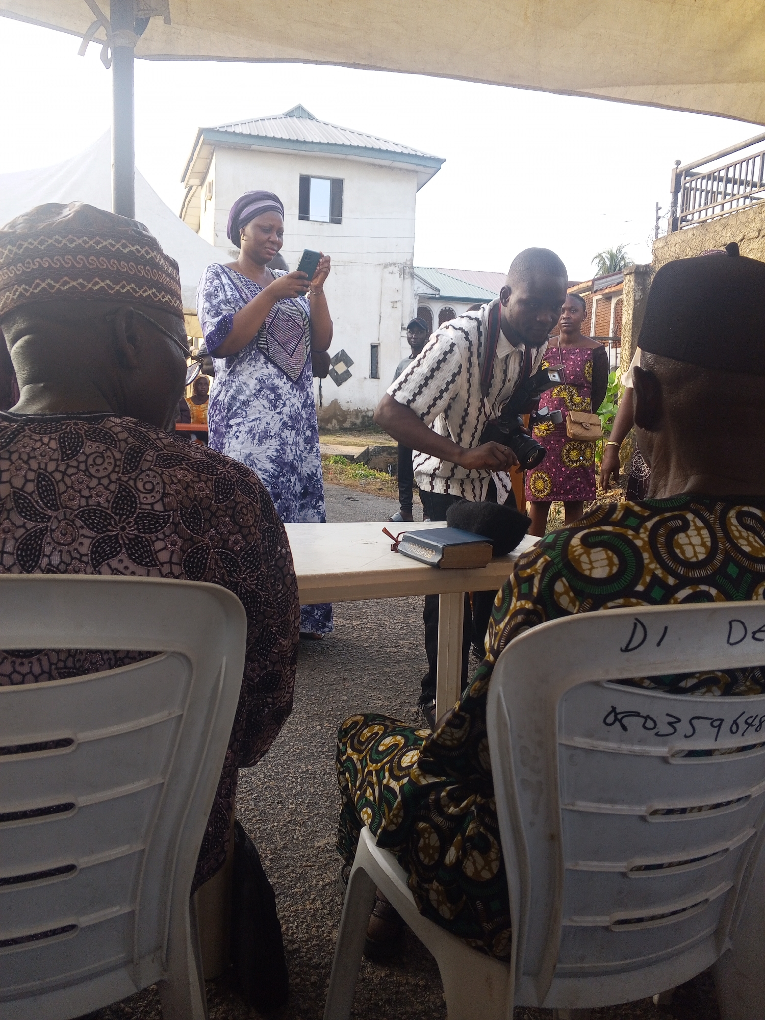 In Lokoja, Nigeria, an outdoor gathering is underway. Three people are visible in the foreground, two men facing away from the camera and a woman in the center. The man on the far left is wearing a traditional cap and a patterned brown garment. The woman in the middle is dressed in a white and blue patterned robe and a purple headscarf, holding a smartphone. The man on the right is wearing a black cap and a patterned green and brown outfit. A white plastic table is in the middle of the frame, with a Bible and a microphone on it. Behind the central figures, a man in a striped shirt is holding a camera with a flash. Another woman in a pink and yellow patterned dress stands behind him. The background shows a white building with a grey roof and a metal fence. The ground is covered in gravel. The lighting suggests it is daytime.