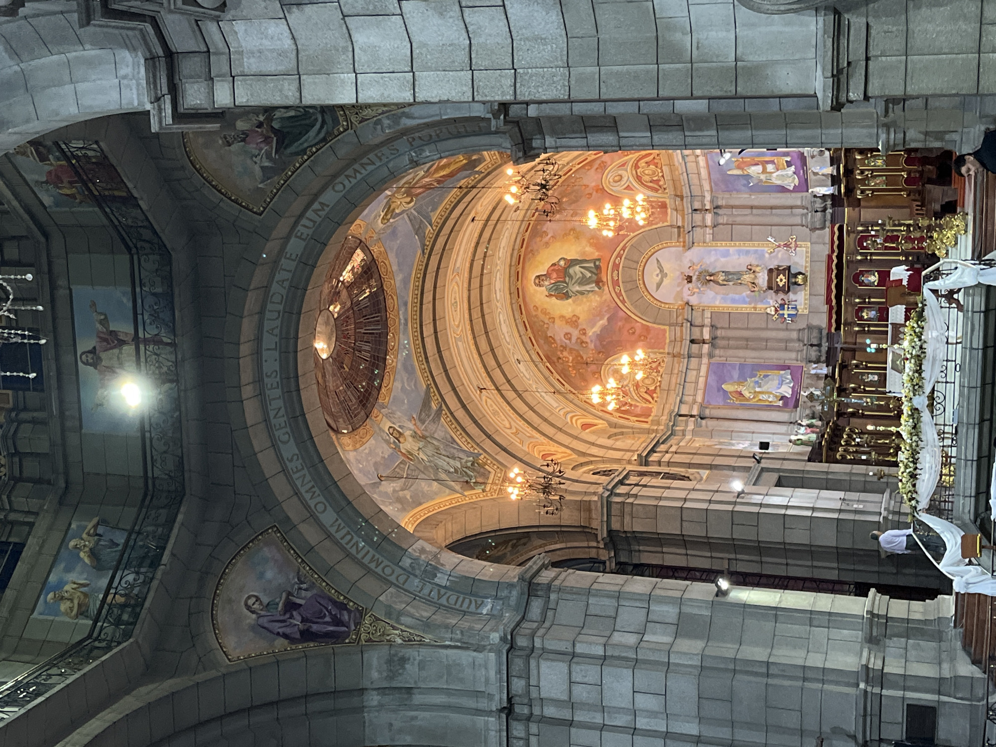 The image shows the interior of the Mérida Cathedral in Mérida, Venezuela, a grand religious building with high arched ceilings adorned with intricate murals and paintings depicting religious figures. Large stone pillars and arches dominate the foreground, framing the view of the altar area which is decorated with floral arrangements and illuminated by ornate chandeliers. Several people are visible, including one individual in a white shirt walking up a staircase on the left, and another person in dark clothing in the lower right corner. The overall scene suggests a place of worship, potentially during a service or preparation for one, given the decorations. The architecture is ornate, characteristic of a historic cathedral, with a blend of stone and painted surfaces.