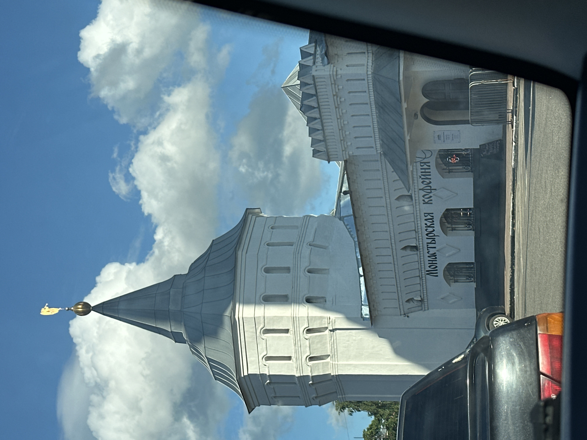 The image shows the Annunciation Cathedral of the Nikolo-Vyazhishchsky Monastery, located in the city of Dzerzhinsky, Russia. The cathedral is a prominent white stone structure with a distinctive octagonal tower topped with a golden angel. The tower has several rows of arched windows. Adjacent to the tower is a lower, white stone building with a serrated parapet and arched entrances. The facade of this building features a sign with Cyrillic lettering that reads "Монастырская кофейня" (Monastery Coffee House). In front of the coffee house, there are metal barriers and some signage. The scene is viewed from inside a vehicle, with the reflection of the car's interior visible in the foreground. A black car is parked on the left side of the frame. The sky is blue with scattered white clouds.