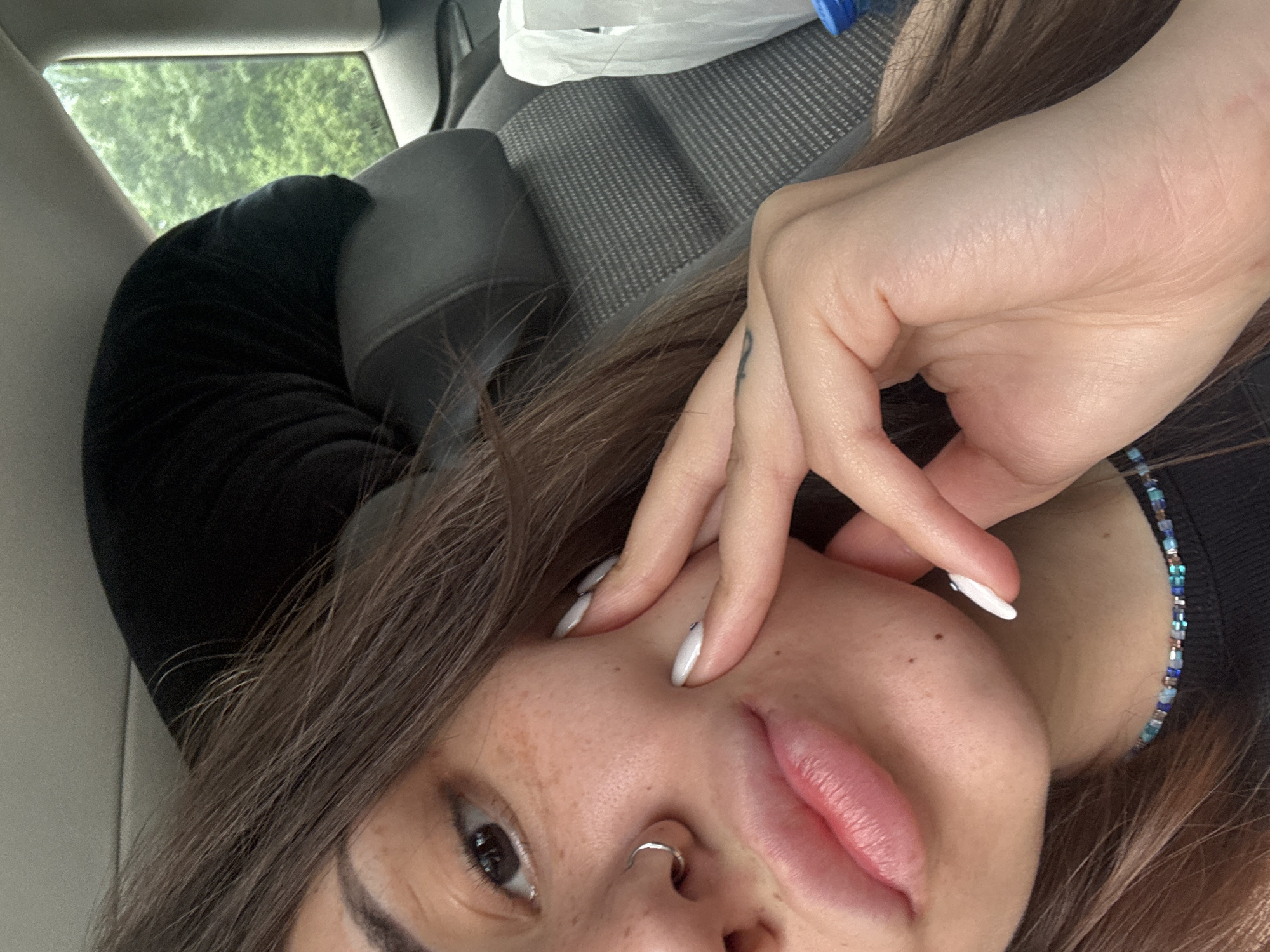 A close-up, angled portrait captures a person, likely female, inside a vehicle in Sofia, Bulgaria. The subject's face, featuring dark brown hair, a visible eye, nose with a silver septum ring, and slightly parted lips, dominates the foreground. Her skin has visible freckles. Her left hand, adorned with white-tipped fingernails and a faint dark tattoo on the back, rests casually near her cheek. A beaded necklace, featuring blue, turquoise, and possibly brown beads, is visible around her neck, complementing a dark garment with a velvet texture on the arm.
The setting is a car interior, with a grey fabric seat and a headrest visible behind the subject. Through the car window, lush green foliage indicates trees or bushes, suggesting the vehicle is either parked or in motion through a natural or treed area. Natural daylight illuminates the scene, implying it is daytime. No text is visible in the image. The overall impression is a casual, intimate selfie taken within a car.