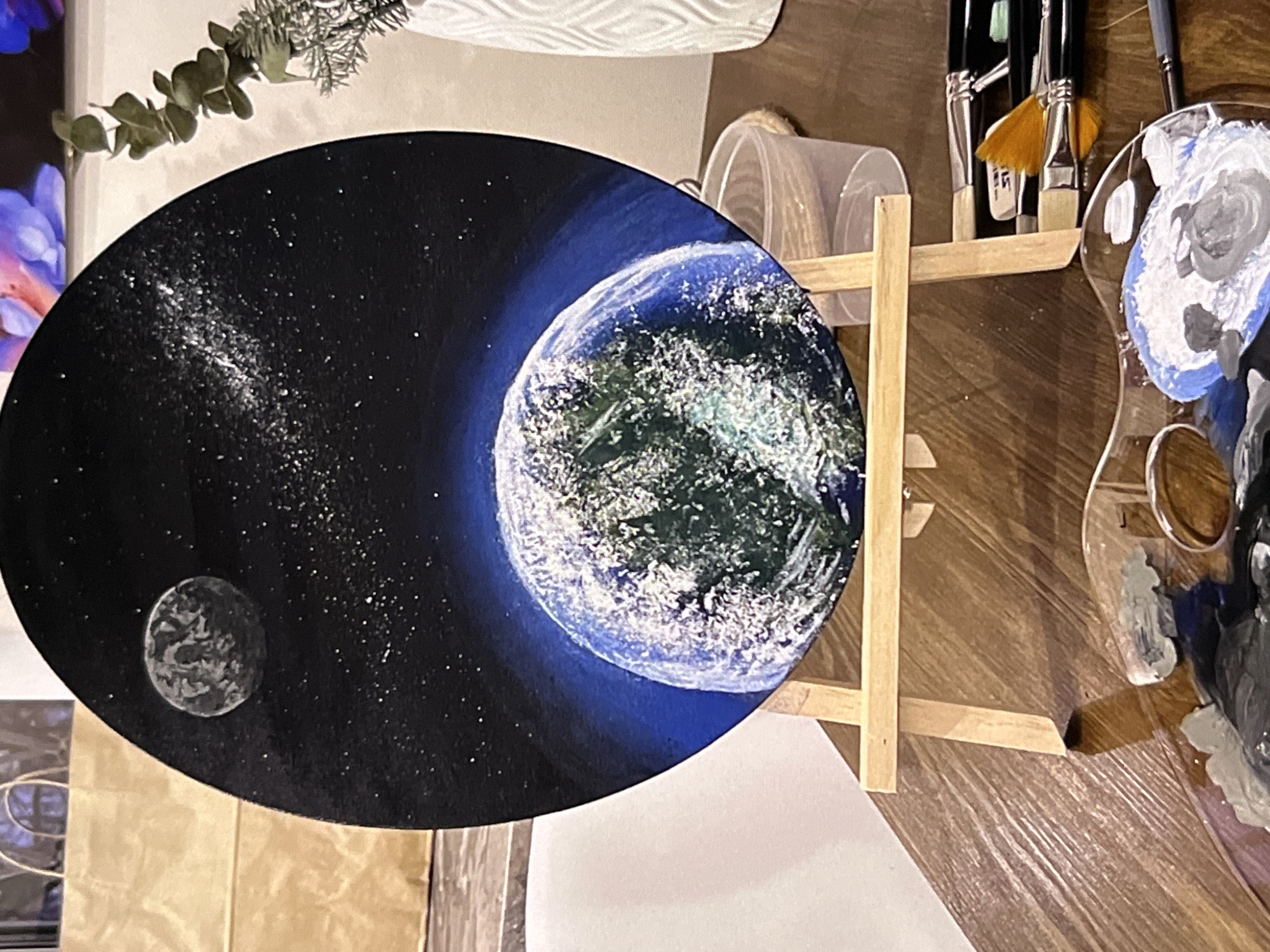 A medium shot depicts a painting of a celestial scene, likely representing Earth and a moon, on a small wooden easel. The painting dominates the foreground. The background shows art supplies, including paint brushes in a container and a paint palette with various colors. The scene appears to be indoors. There are no people visible in the image. The location is unknown, Indonesia.