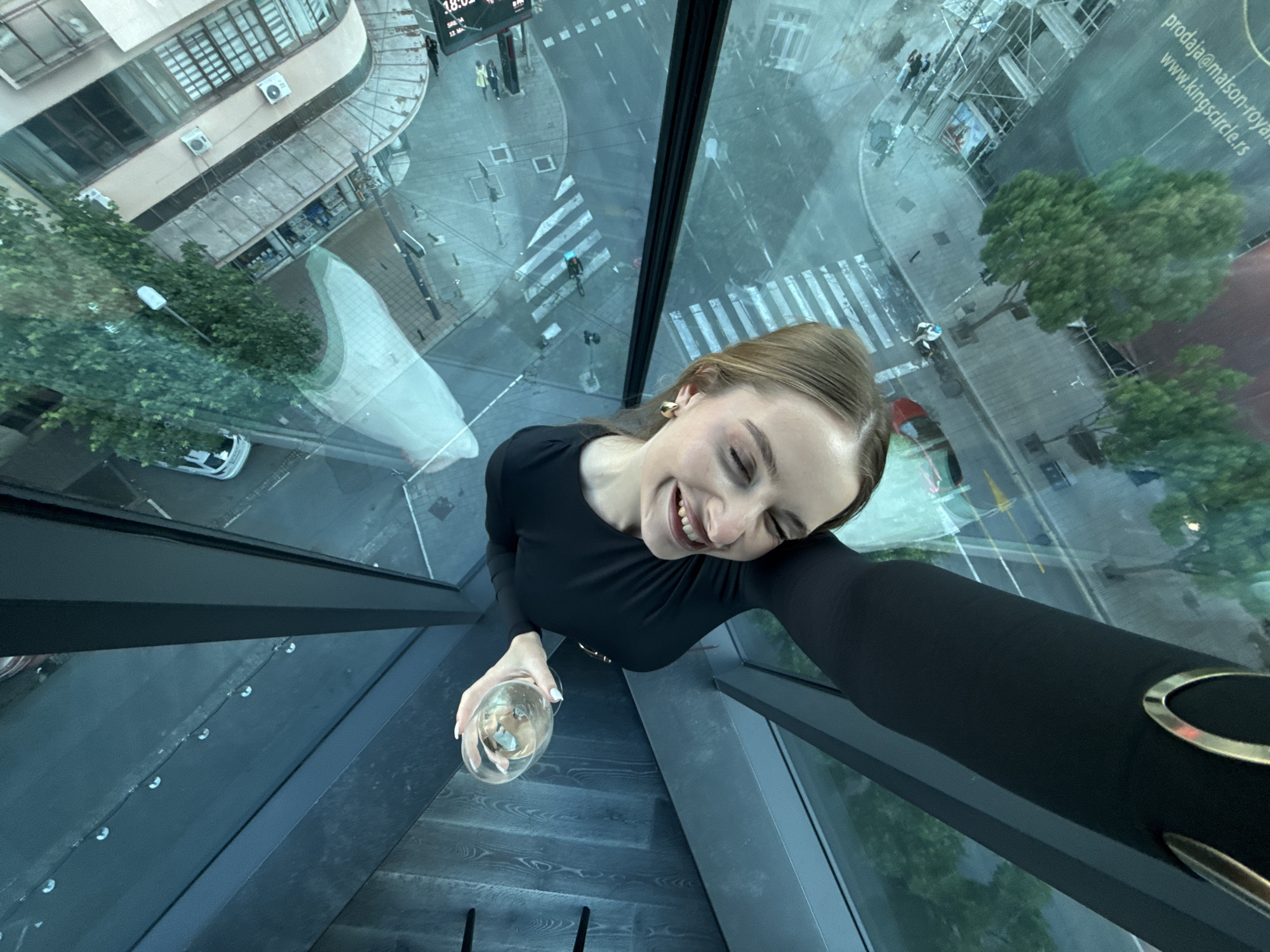 A high-angle selfie shows a woman holding a glass of liquid, likely wine, standing on a glass-floored platform overlooking the city of Belgrade, Vracar Urban Municipality, Serbia. The woman, dressed in black, smiles into the camera. Below her is a view of a street with pedestrian crosswalks, trees, cars, scaffolding, and buildings. There is a digital time display on a building in the distance. Text is visible on another building that says "prodaja maison-royal www.kingscircle.rs". The photo is taken from a high vantage point, perhaps a skyscraper.