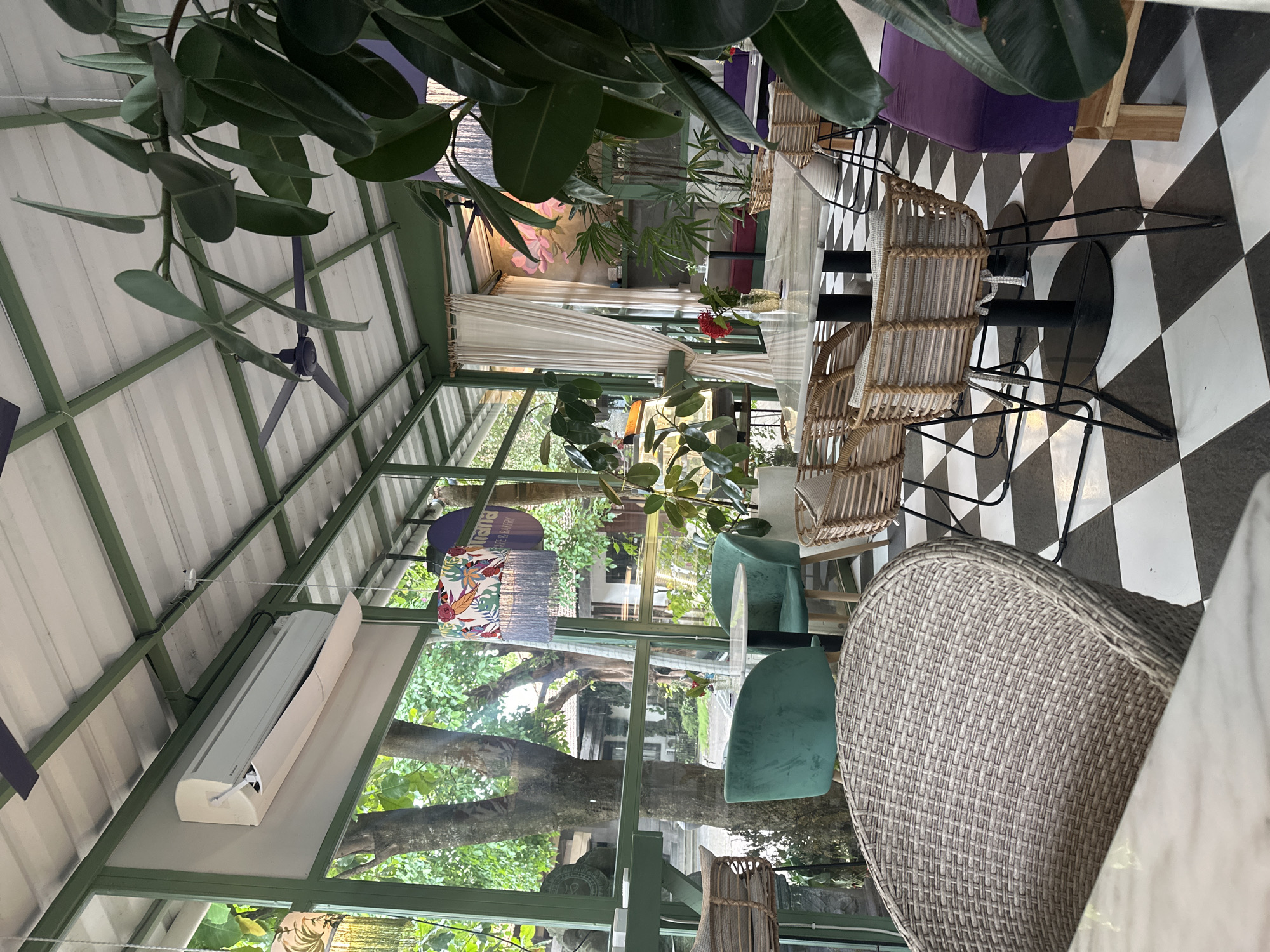 This image shows an interior scene, likely a restaurant or cafe in Celuk, Indonesia. The space has a greenhouse-like structure with a white ceiling, green metal framework, and large windows. Outside the windows, lush green trees and foliage are visible. Inside, a table with a white marble top is surrounded by wicker and plush chairs. The floor is covered in a black and white checkered pattern. A ceiling fan is visible in the upper left of the frame. The scene is well-lit with natural light streaming through the windows.