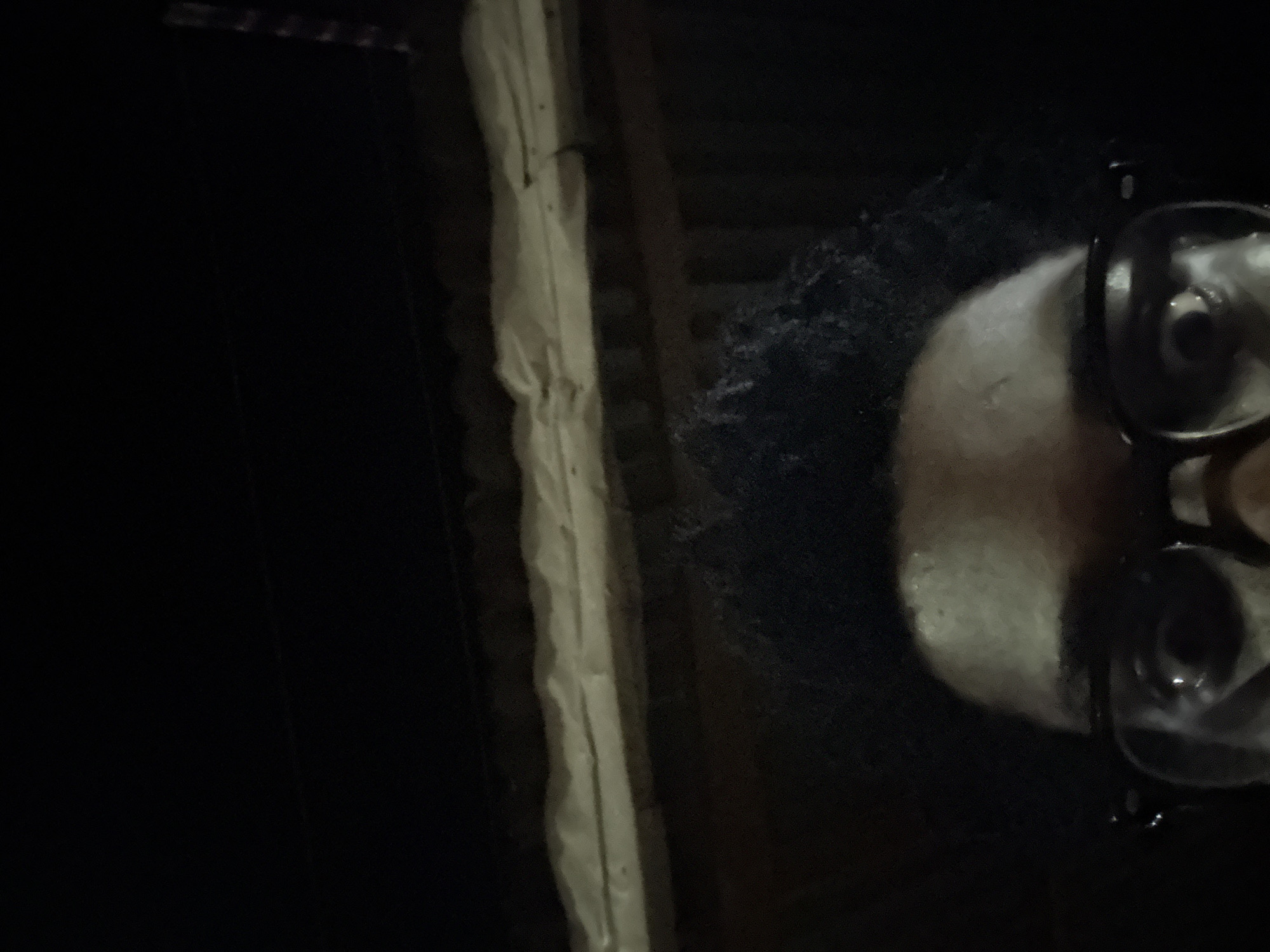 The image shows a dimly lit scene, likely indoors, featuring a vertical white or light-colored beam against a dark background. Adjacent to the beam is a blurry, seemingly organic form, possibly an animal or a piece of food, with reflective, circular objects that could be glasses or lenses nearby. The background is largely obscured, but suggests a structure or enclosure of some sort. The location given is Ihiagwa, Nigeria, but there's no immediately discernible evidence within the image to confirm this location specifically.