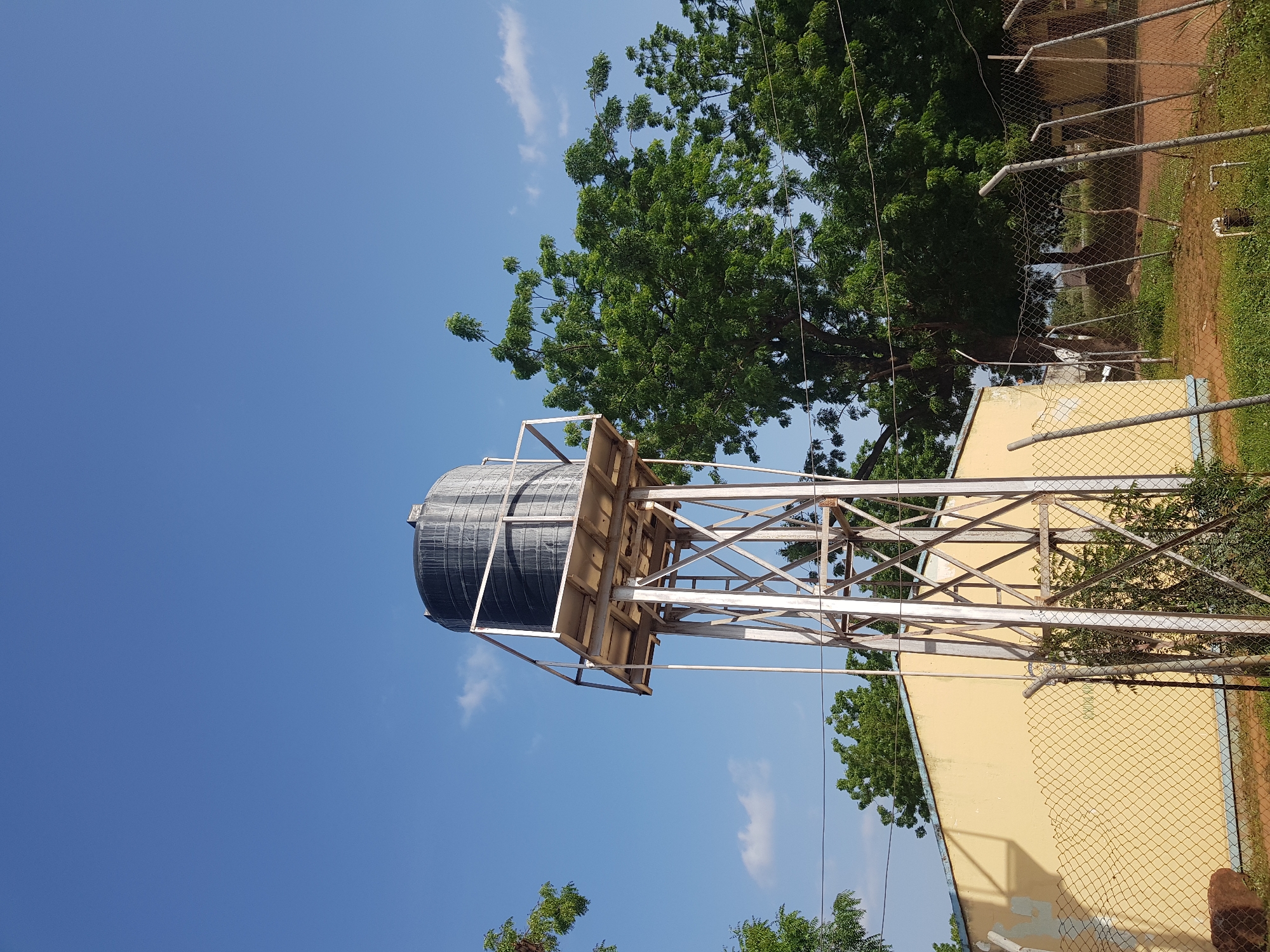A black cylindrical water tank is elevated on a metal support structure under a clear blue sky with sparse white clouds. The water tank is positioned on the right side of the frame, with its base on a square metal platform. The supporting structure is made of interwoven metal beams. To the left of the water tank and support structure is a pale yellow building with a blue trim. A chain-link fence runs in front of the building, and beyond that is dry, patchy grass and more trees. The scene appears to be an outdoor area, possibly in a rural or semi-urban setting in Jimeta, Nigeria, characterized by the presence of vegetation and a utilitarian water storage system. There are no people visible in the image.