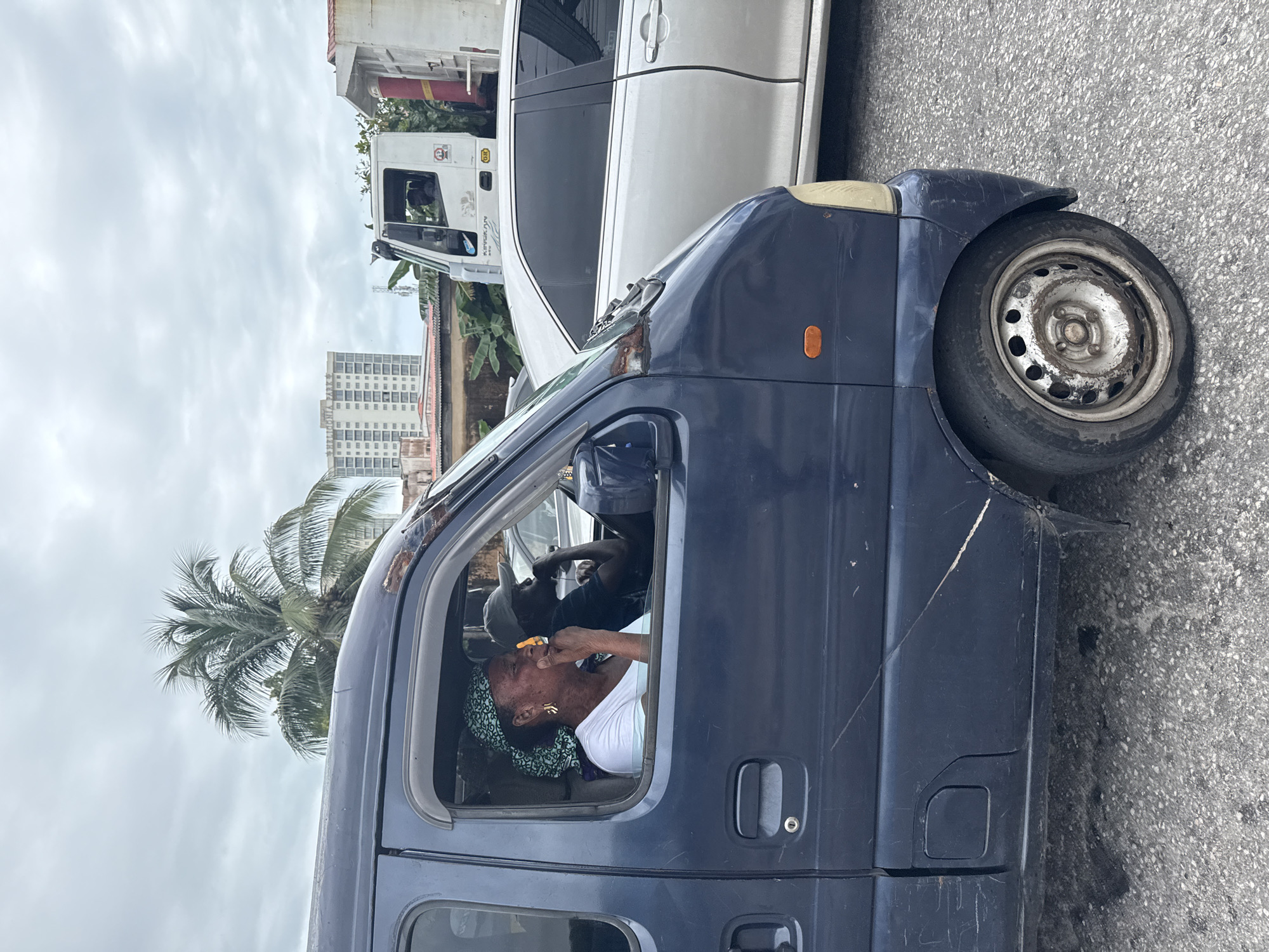 The image shows a woman in the driver's seat of a dark blue minivan. She is wearing a white t-shirt and a green and white patterned headscarf. She has earrings and appears to be putting something to her mouth with her right hand. Another person, a man wearing a grey baseball cap and a dark t-shirt, is visible in the passenger seat behind her.
The minivan is parked on a paved road, with a white truck and a white car behind it. A tall building and palm trees are visible in the background, suggesting an urban setting in a tropical or sub-tropical location. The sky is overcast, indicating it might be a cloudy day. The overall scene appears to be a street in Ikoyi, Nigeria. The van has visible wear and tear, with scratches and rust on the exterior. The wheel of the minivan is also visible, appearing somewhat dirty. There is no discernible text in the image.