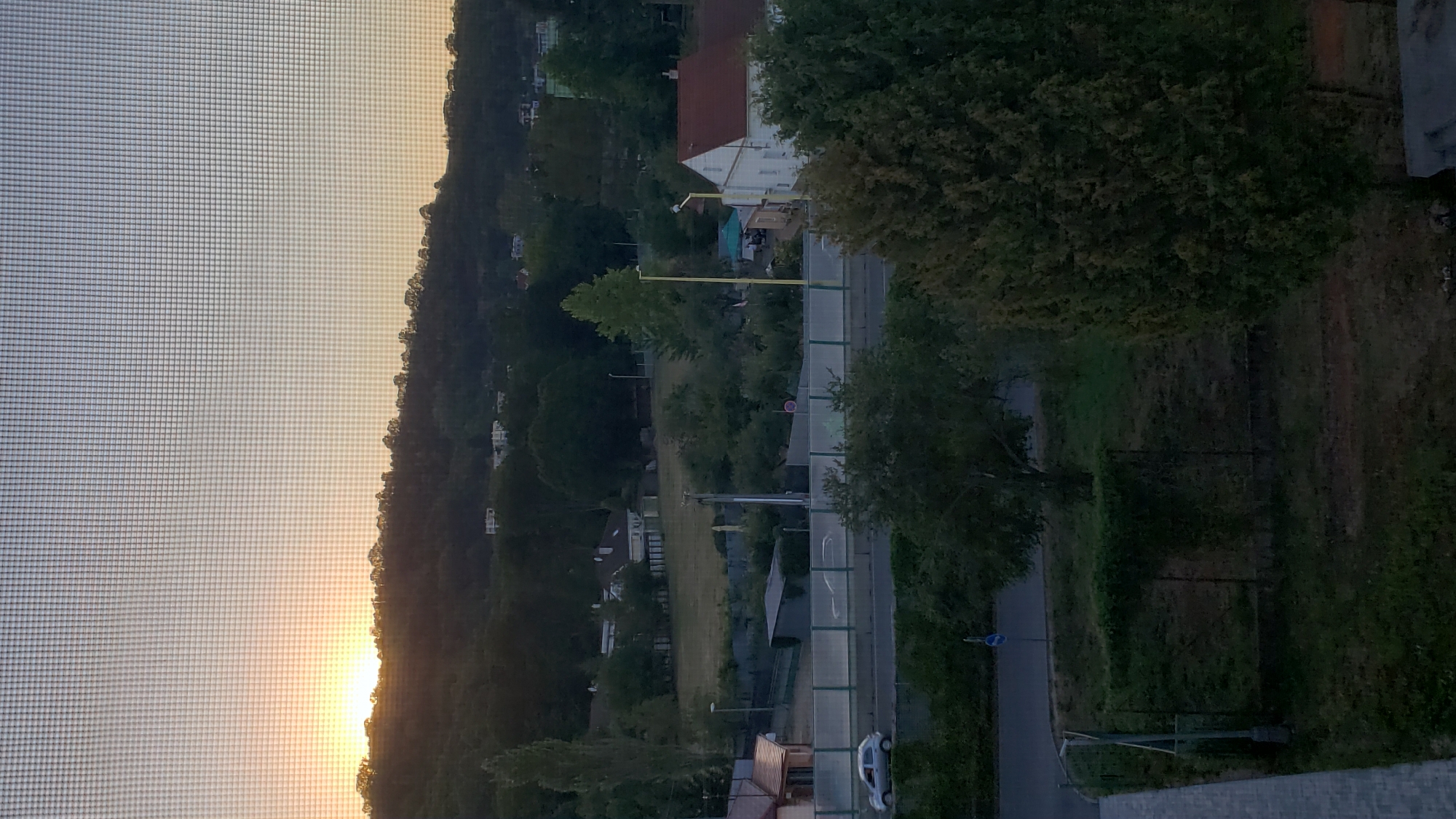The image depicts a view through a window screen of a residential area in Prešov, Slovakia, during either sunrise or sunset. A silhouetted treeline borders a gently sloping hill in the background, with the sun peeking over it on the left side of the frame. Below the hill, several houses and buildings are visible amidst trees. A road with a light-colored barrier runs across the middle ground, and a silver car is seen on the road. In the foreground, a paved walkway or balcony is partially visible on the left, with a grassy area and trees to the right. The overall scene is characterized by a soft, ambient light and a peaceful, suburban atmosphere.