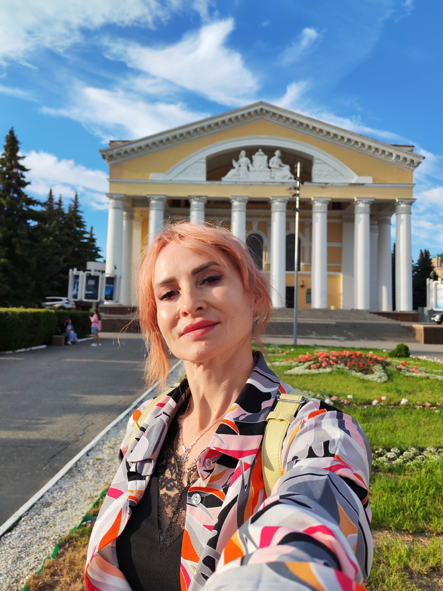 A woman with pink hair takes a selfie in front of the National Youth Theater of the Republic of Mari El in Yoshkar-Ola, Russia. The theater is a large, yellow, neoclassical building with white columns and a triangular pediment. A flower bed with red and white flowers is in front of the theater, and trees line the sidewalk. The sky is blue with white clouds. The woman is wearing a colorful, patterned jacket and a black lace top. She is smiling at the camera.