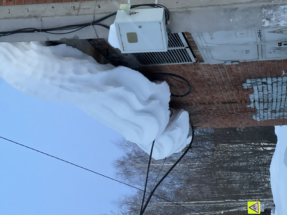 The scene, set in Noviy, Russia, depicts a severe winter environment. A massive, undulating accumulation of white snow, exhibiting a layered, wave-like texture, heavily overhangs the roofline of a red-brick building. This snow cornice rests on or is in close proximity to several black utility cables strung along the building's facade. To the right, a concrete utility pole supports multiple black electrical wires and a white rectangular electrical box, which has a thin layer of snow on its top surface. The red-brick wall below the snow displays weathered sections and some light-colored paint or graffiti. Beneath the overhanging snow, a horizontal slatted vent is visible, and below it are two light-colored metal utility doors marked with Cyrillic text, including "ТП-Р2" and "ТП-1." The ground in the foreground is covered in deep snow. In the lower-left, a yellow triangular road sign with a black border indicates a children's crossing, supplemented by a smaller rectangular sign displaying "100m." Bare deciduous trees are visible in the distant background against a clear, light blue sky. No individuals are present in the image.