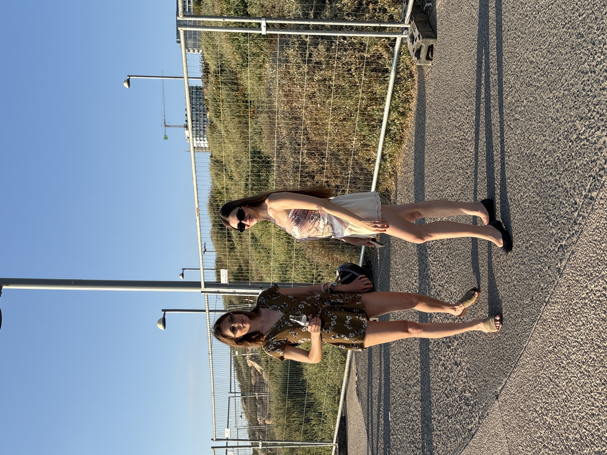 Two women stand on a paved surface in front of a wire fence. The woman on the left wears a floral print romper and sandals and holds a bottle. The woman on the right wears a strapless top and a light-colored skirt and flats. Both women are wearing sunglasses. The background shows a construction crane, buildings, and vegetation under a clear blue sky. The scene appears to be in Tel Aviv, Israel, potentially near a beach or coastal area, given the fence and the construction.
