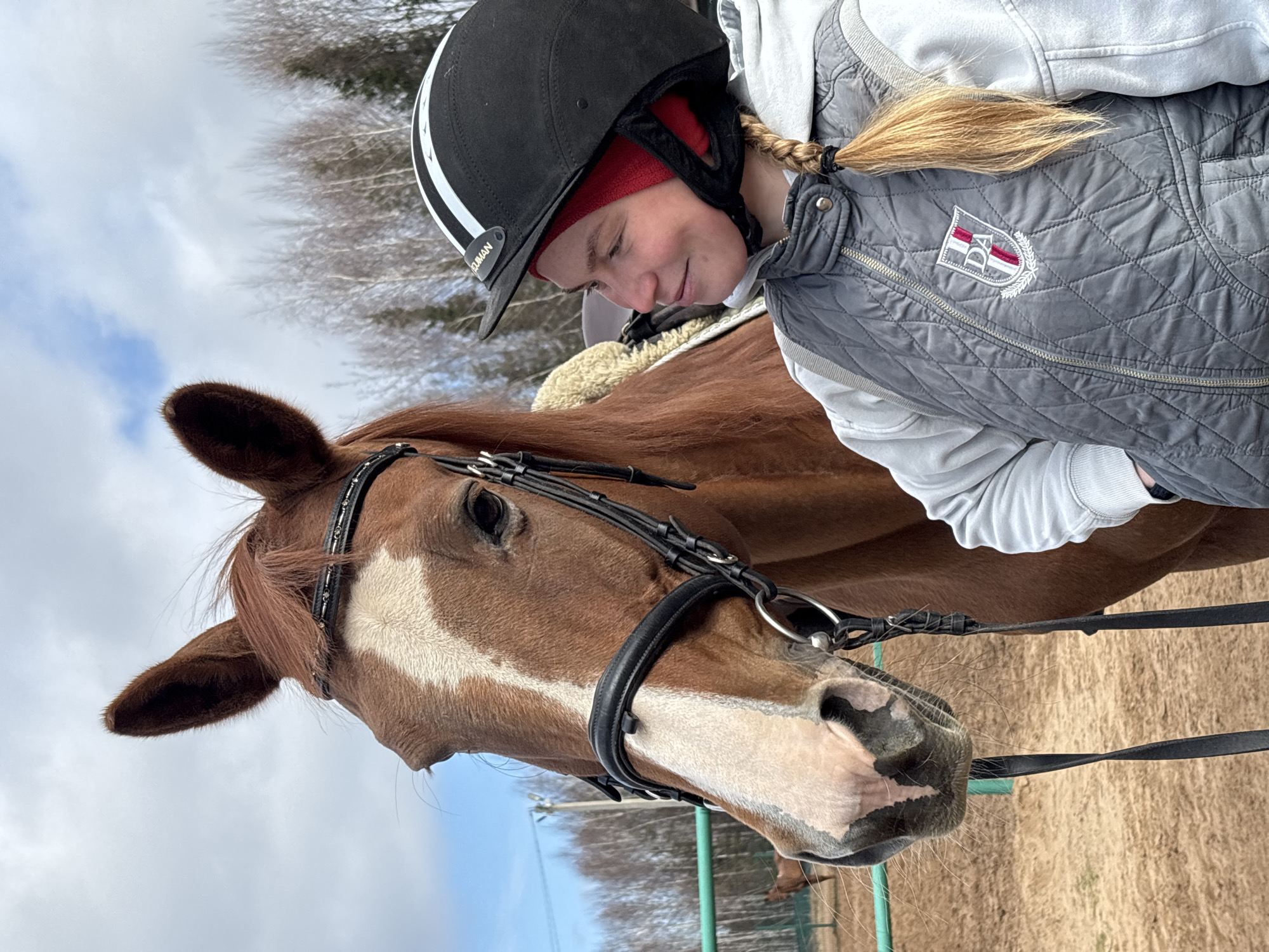 A young woman wearing a black riding helmet, red headband, gray vest, and white hoodie is standing next to a chestnut horse with a white blaze. The horse is wearing a bridle. The background includes trees and a partly cloudy sky. The location appears to be an outdoor riding area.