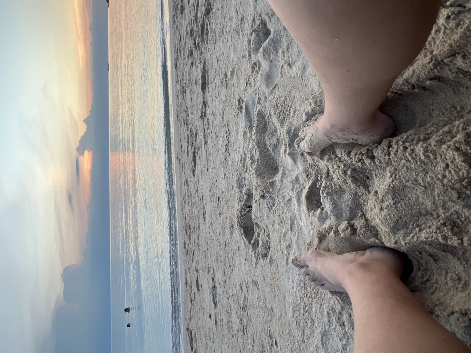 A first-person perspective shot captures feet partially buried in damp sand on a beach in Ko Pha-ngan District, Thailand. The sun is setting over a calm ocean, casting a warm, pastel glow across the sky and water. Two individuals are visible swimming in the distance. The sand appears wet and textured, with footprints and slight indentations indicating recent activity. The waves are gentle, lapping at the shoreline. The overall scene conveys a sense of tranquility and relaxation at the end of the day.