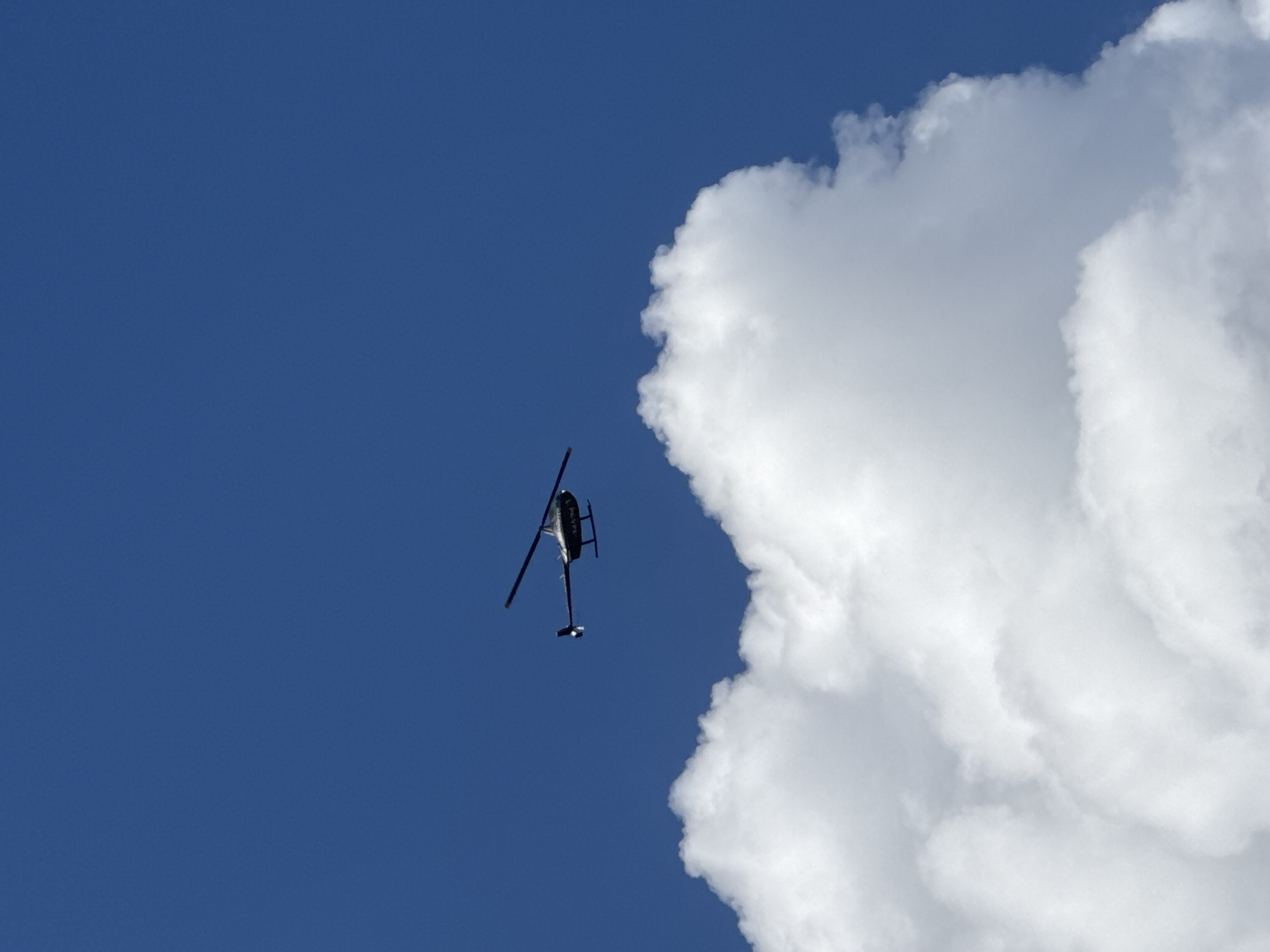 A dark-colored helicopter is captured mid-flight against a clear blue sky. The helicopter is positioned in the upper right quadrant of the frame, its rotors spinning. Below the helicopter, a large, fluffy white cloud dominates the lower portion of the image. The scene appears to be an aerial view, likely taken during daytime due to the bright sky. There are no people visible in the image. The location in Kuta Selatan, Indonesia, is not discernible from the visual information provided in the image.