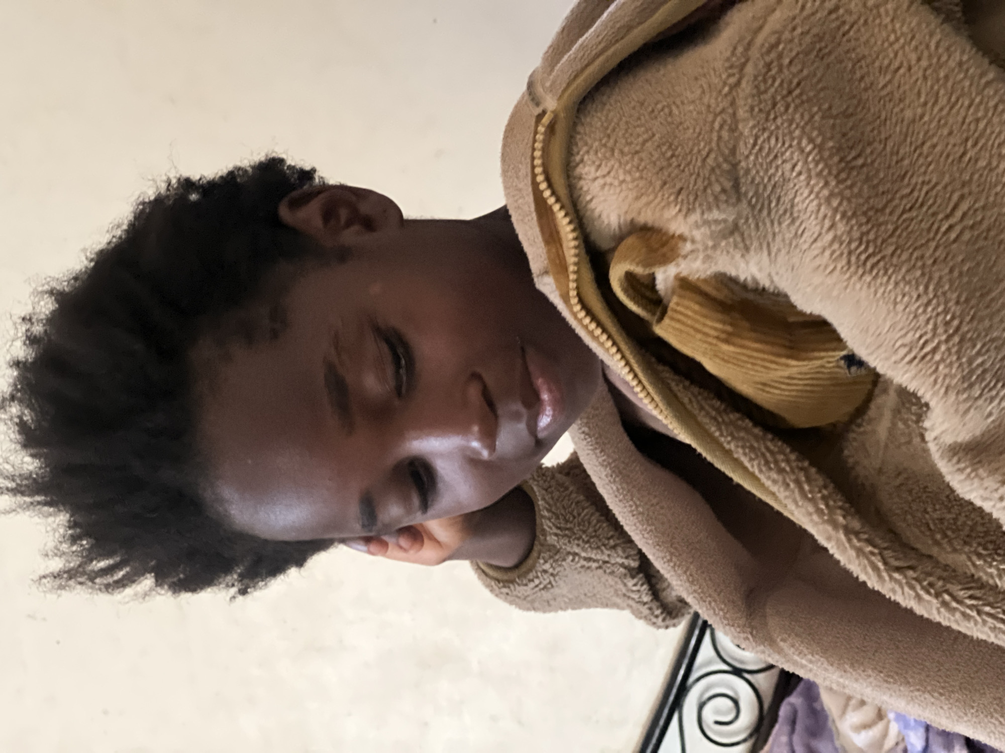 The image is a medium shot of a young Black person, likely a child, with dark, curly hair styled upwards. They are wearing a tan, fuzzy zip-up jacket. The person is looking down and to their right, with their right hand resting on their head and ear. The background appears to be a plain, lightly colored wall. There are no discernible elements in the image that would allow for the identification of a specific location within the city of Jos, Nigeria.