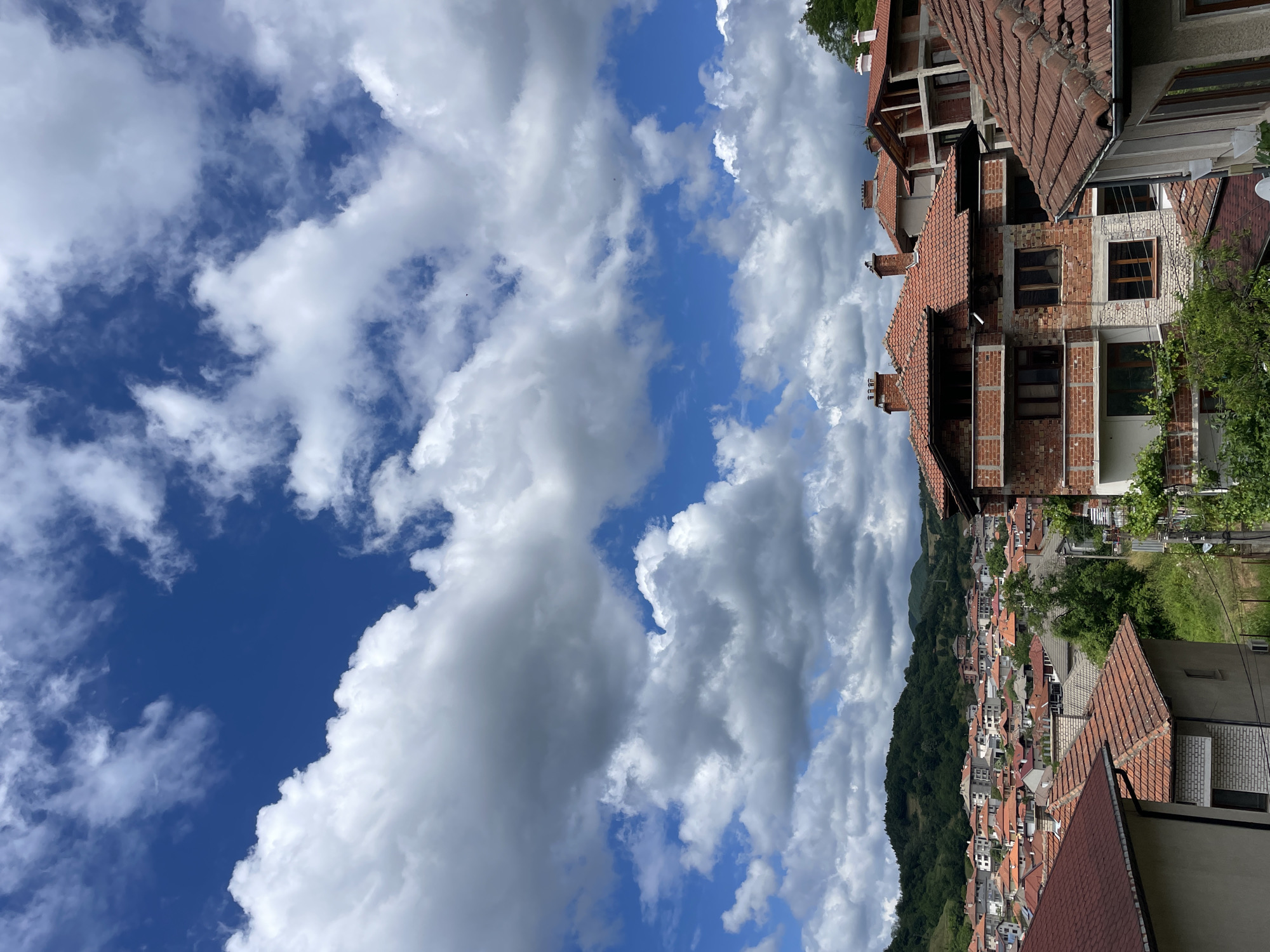 This is a daylight, eye-level, medium-shot photograph taken outdoors in Zlatograd, Bulgaria. The image captures a view of a residential area with houses, rooftops, and a hillside in the background under a partly cloudy sky.
In the foreground and middle ground, several houses with red tiled roofs are visible. One prominent building in the middle ground is under construction, with exposed brickwork and balconies. Another house in the lower right corner also features a red tiled roof and part of its facade.
The background shows a densely populated residential area with buildings of various sizes and architectural styles, many also topped with red tiles. The houses extend up a gently sloping hillside covered with green trees and vegetation. The sky above is a vibrant blue, dotted with numerous white, fluffy cumulus clouds.
There are no people or animals visible in the image. The scene depicts a typical urban or semi-urban setting within a hilly landscape, characteristic of Zlatograd.