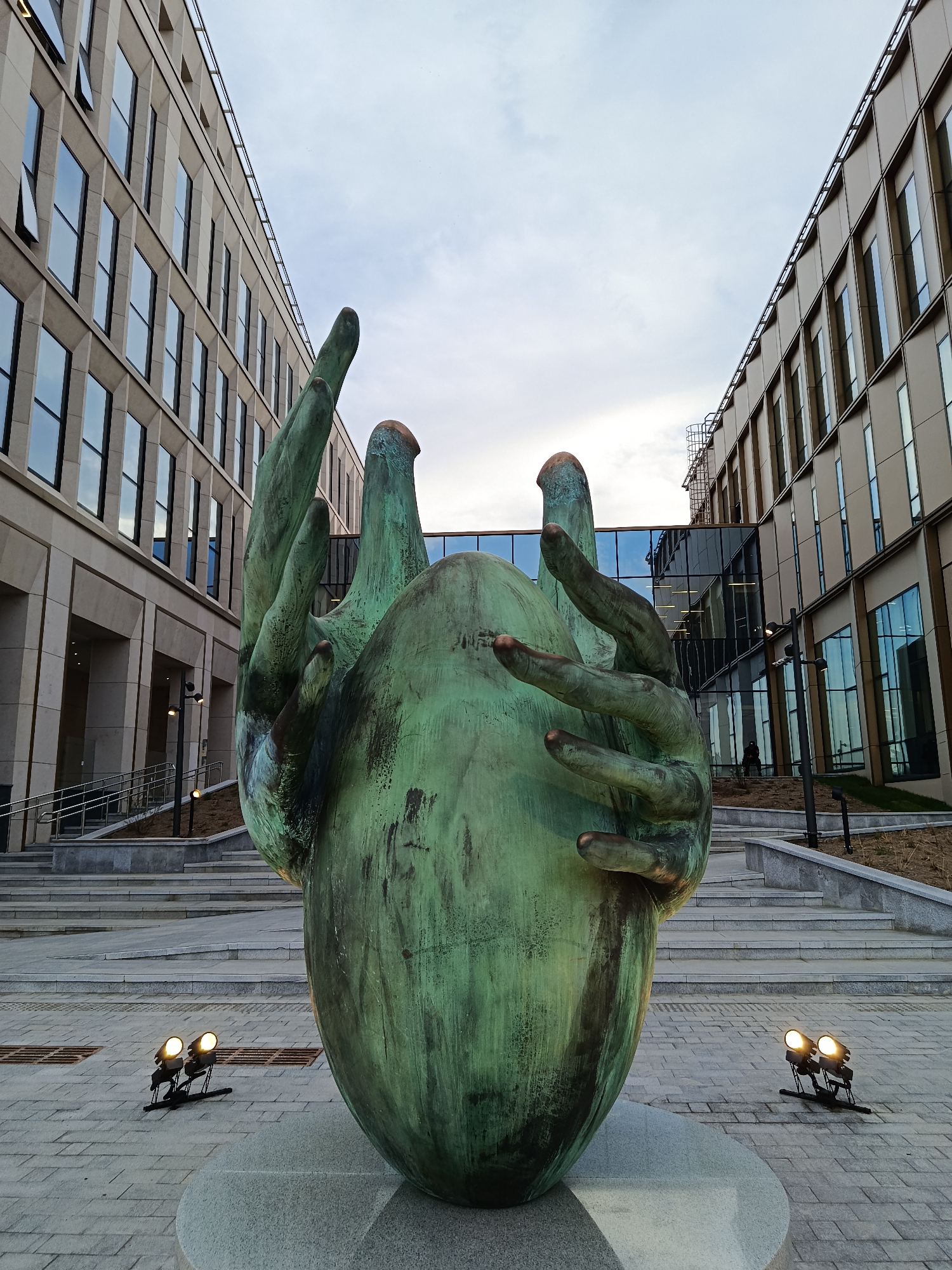 This is an eye-level shot of a bronze sculpture of two hands holding a green, egg-shaped object in front of two modern buildings in Moscow, Russia. The sculpture is situated on a circular stone base and is illuminated by two sets of spotlights on either side. The buildings on either side of the sculpture have multiple stories and uniform window patterns. The sky above is overcast. The foreground is a paved area. Stairs lead up to the building on either side of the sculpture.