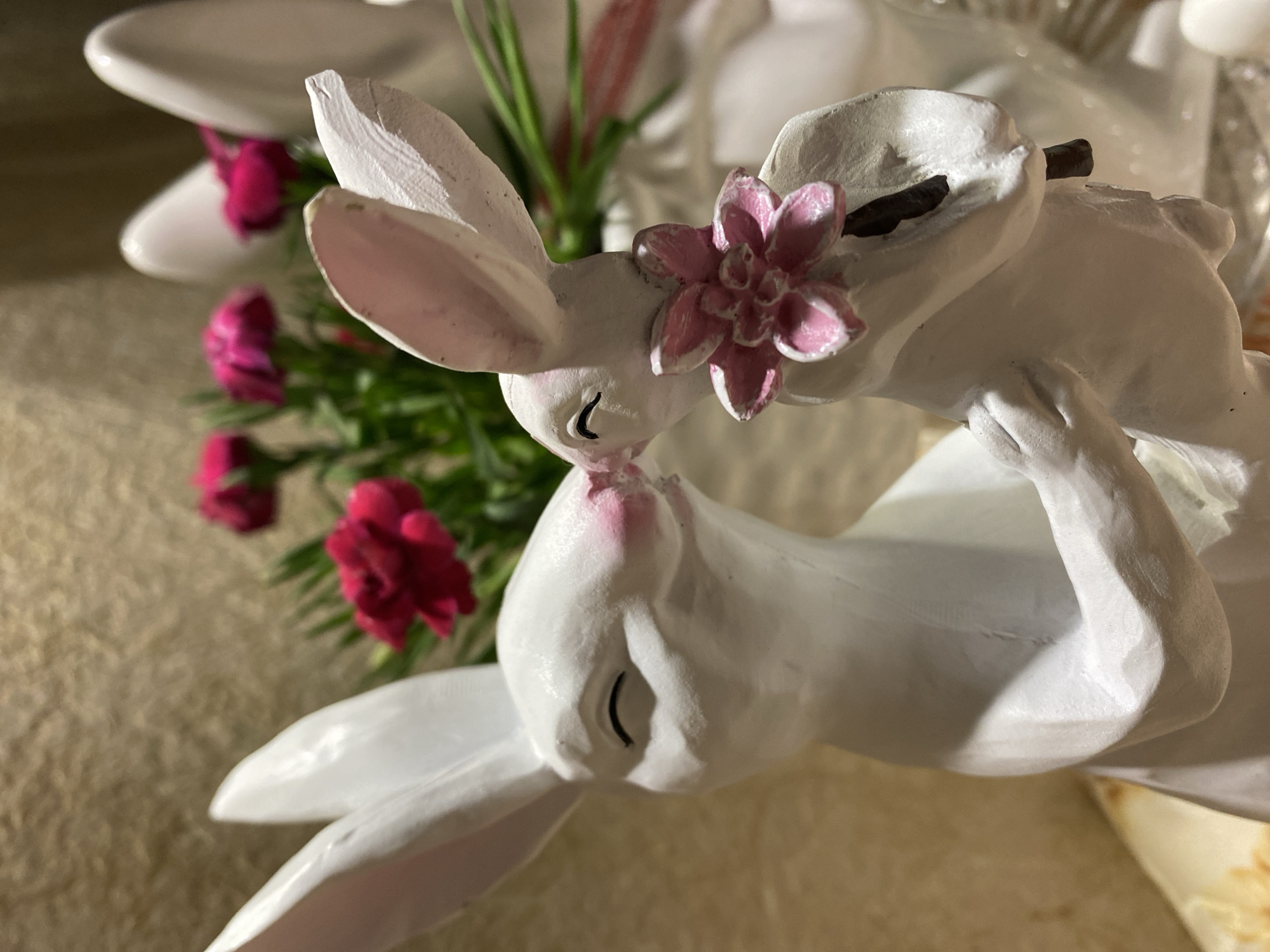 A close-up shot features a pair of white ceramic bunnies posed nose-to-nose, seemingly kissing. The bunny on top has a pink flower adorned on its head. In the background, there's a blurry arrangement of dark pink carnations and green foliage. The bunnies are positioned on a tan, textured surface. The setting is indoors, with a few other indistinct objects visible in the far background. The image does not depict Kryvyi Rih, Ukraine, nor contain any details that would suggest its location.
