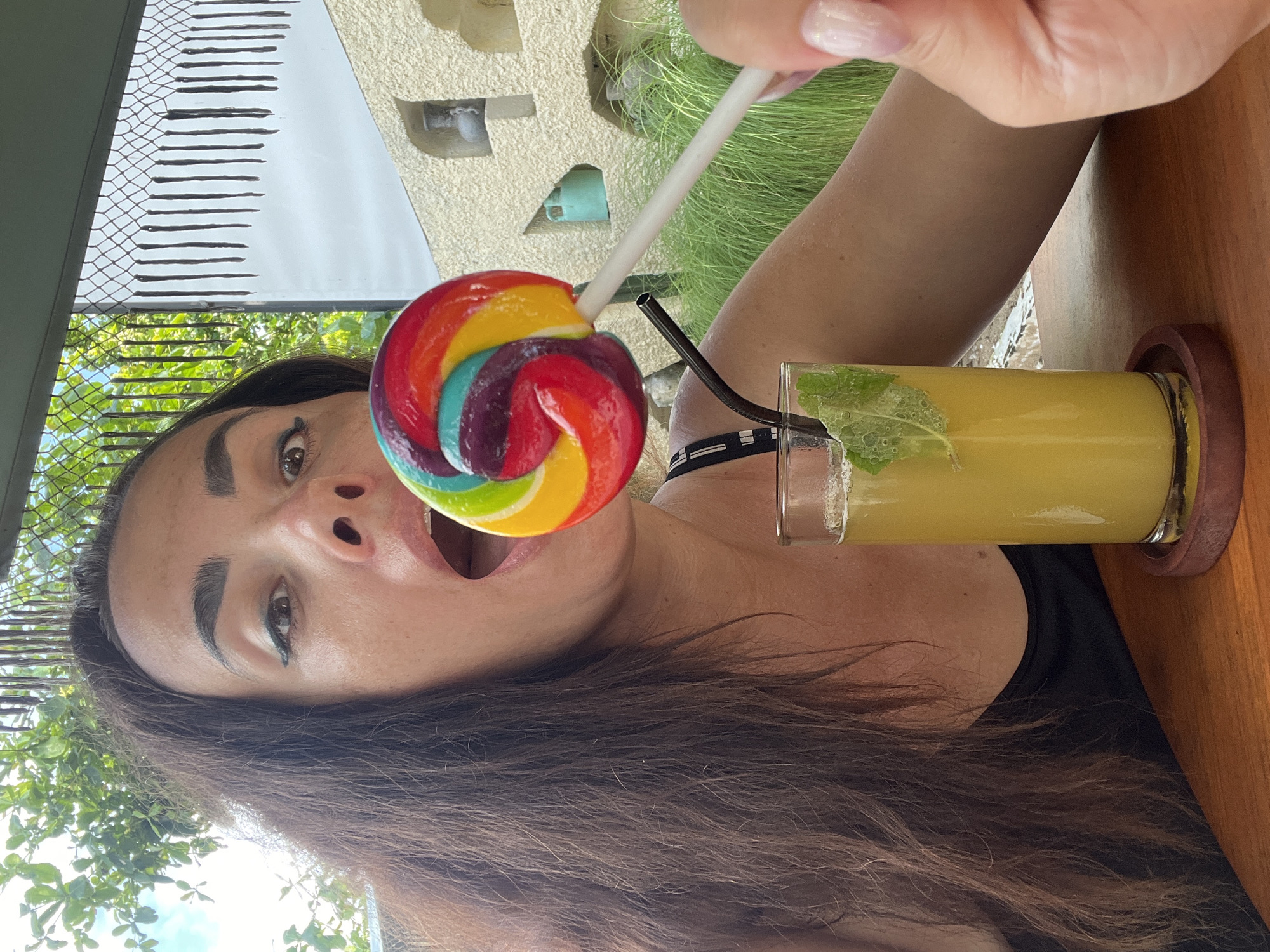 A medium shot captures a woman in Kuta Selatan, Indonesia, leaning forward with her mouth open towards a rainbow-swirled lollipop she holds with her right hand. Her dark brown hair cascades down her shoulders, framing her face. She wears a black top. Beside her, a tall glass filled with a yellow beverage and mint leaves is placed on a wooden coaster. The background features lush green foliage, a decorative fence with vertical black strands, and a beige stone wall with niches. The lighting suggests an outdoor daytime setting.