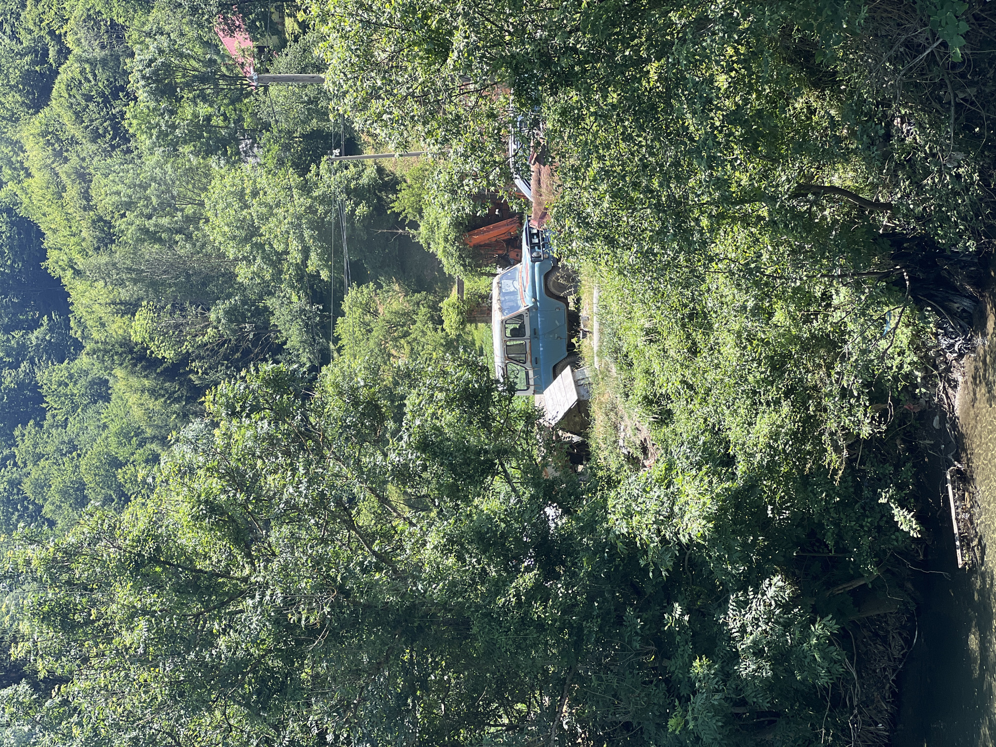 This image depicts a scene in Moračko Trebaljevo, Montenegro, characterized by dense green foliage and a body of water in the foreground. A vintage blue SUV is parked on an incline amidst overgrown vegetation. Behind the SUV, an orange piece of construction equipment, possibly an excavator, is visible. In the distance, a red-roofed house is nestled among trees on a hillside. The water in the foreground appears shallow, with debris floating on its surface. The overall impression is one of a rural, somewhat neglected setting.