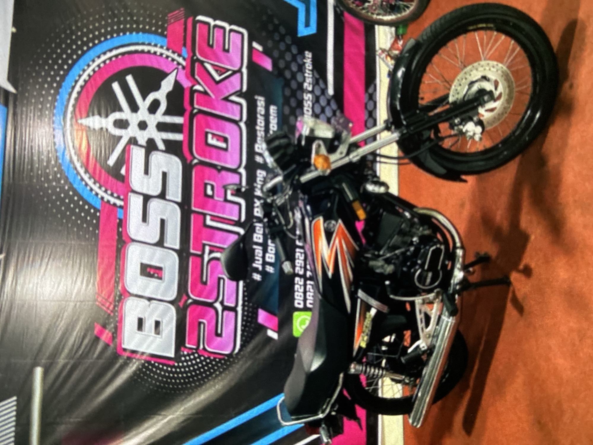 The image depicts a black motorcycle positioned in front of a backdrop with the words "BOSS 2STROKE" prominently displayed. The backdrop features the Yamaha logo in pink and blue, along with text in Indonesian, possibly related to buying, selling, and restoring PX King motorcycles. A phone number is also present on the backdrop. The location is likely Lhokseumawe, Indonesia, based on the language used on the backdrop and the context of the objects within the frame. The motorcycle is the main subject, with the backdrop serving as a promotional or advertising element.