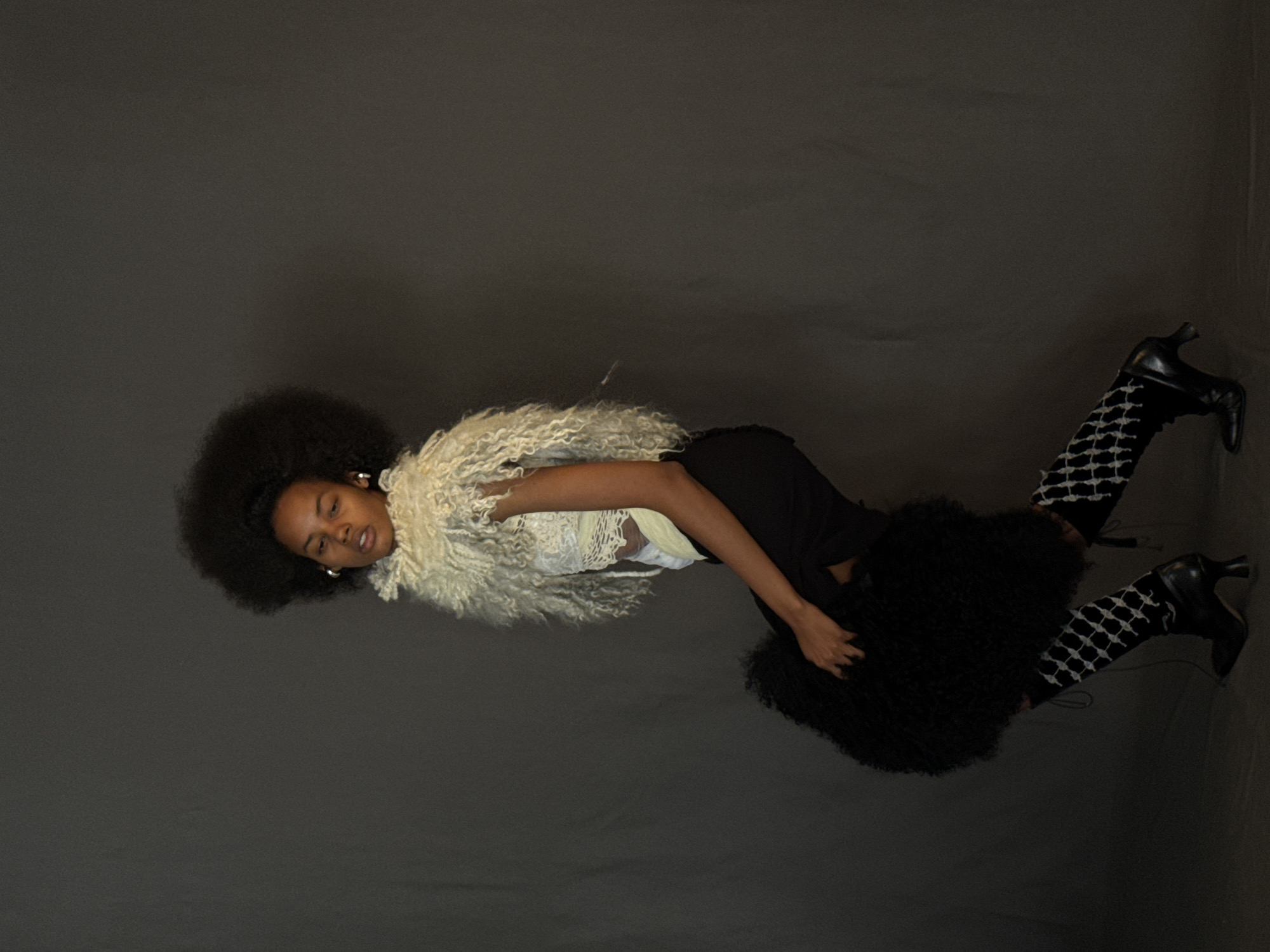 A woman with dark skin and a large afro lies on a gray surface. She is wearing a cream-colored curly shawl, a light-colored lace top, a black skirt, patterned tights, and black heeled boots. Her right hand rests on her skirt. The location and city are not discernible.