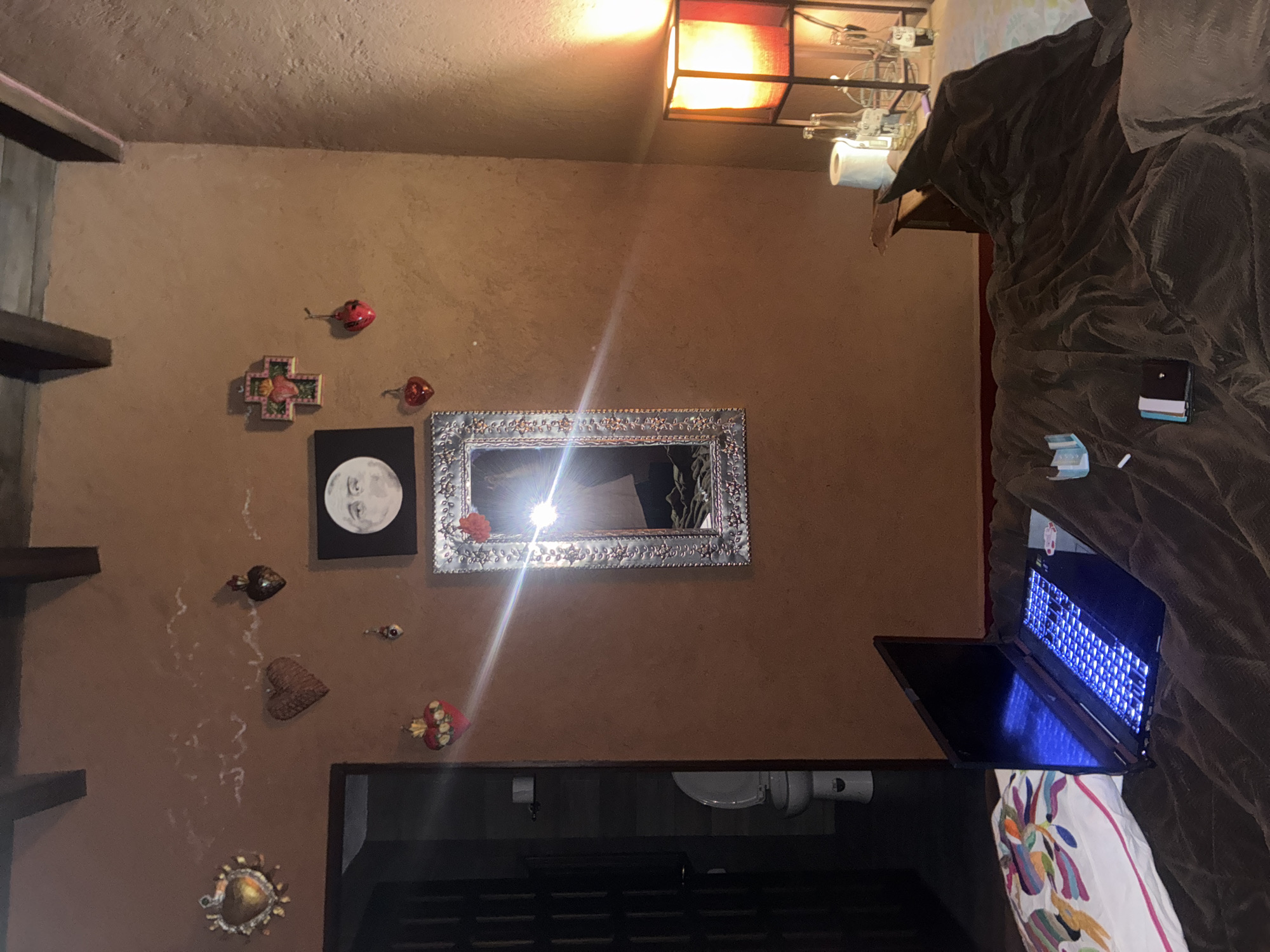 The image shows a room in what appears to be a home. On the left wall are wooden shelves and various decorative items, including a cross, moon art, hearts, and a sun. A framed mirror hangs on the wall. Below the wall decorations is a dark surface that appears to be the top of a cabinet or console. A toilet and sink are positioned below the mirror and cabinet. A laptop is open and illuminated on the right side of the image, resting on bedding and pillows. A dark blanket covers the bed. The room is lit by a square lamp on the ceiling. The specific location in the city of Cañada de Madero, Mexico, cannot be determined based solely on this image. No people are visible.