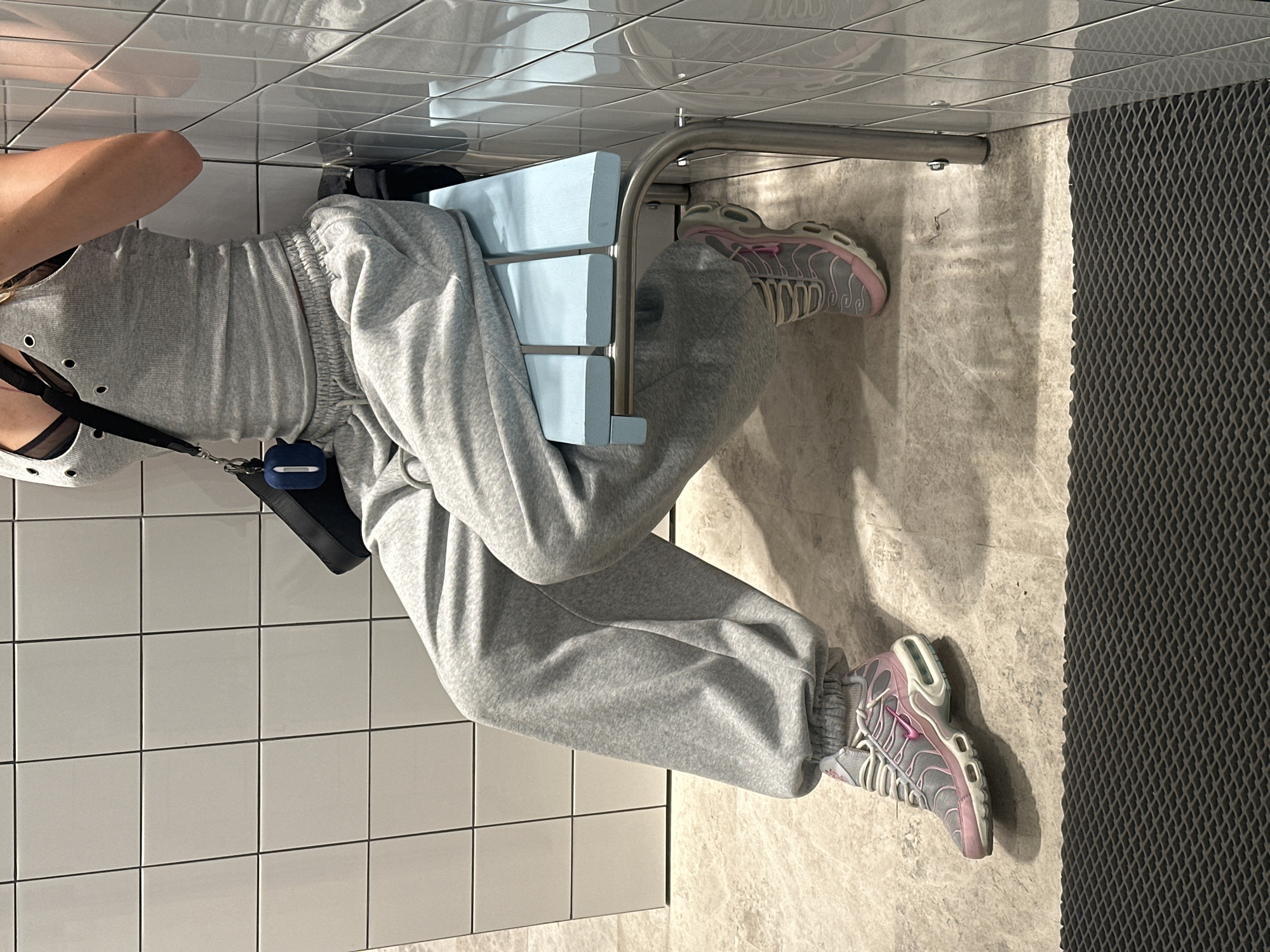 The image shows a person sitting on a blue slatted bench in what appears to be a public restroom or changing area. The walls are tiled with white square tiles. The person is wearing grey sweatpants, a grey tank top with metal grommets, and grey and pink athletic shoes. A black purse with a blue AirPods case is slung over the person's shoulder. The floor is made of marble-like tiles, and a black textured mat is visible in the foreground. The scene is well-lit, with shadows cast on the floor. There is no specific indicator of the location being Moscow, Russia; the description is based solely on the visual elements present.