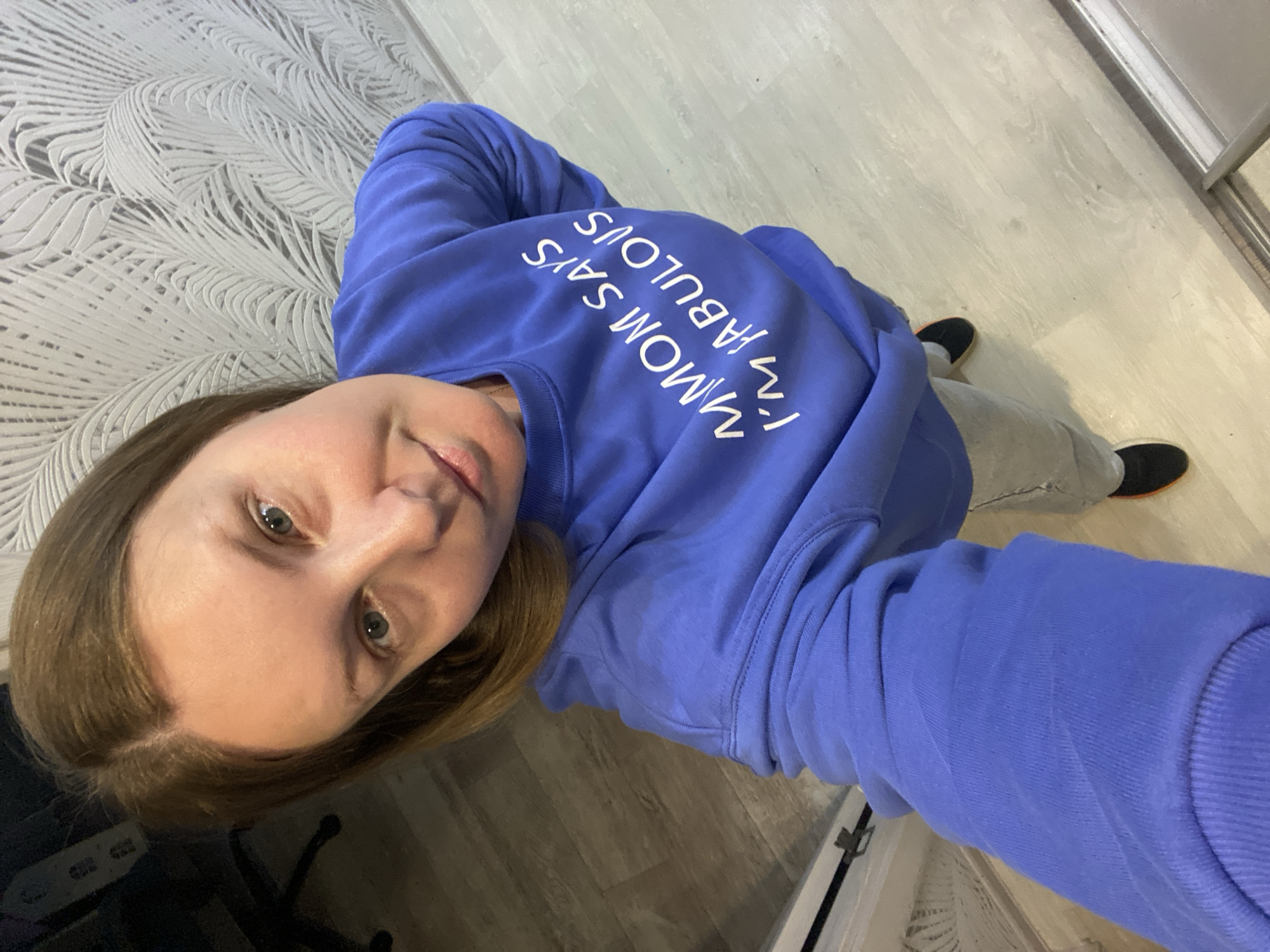 A woman is taking a selfie indoors. She is wearing a blue sweatshirt with the text "MOM SAYS I'M FABULOUS" printed on it. She is also wearing gray sweatpants and black shoes with orange soles. The floor is made of light-colored planks. In the background, there is wallpaper with a leaf pattern on one wall and a white closet with a silver handle on another wall. The location is likely in Noviy, Russia.