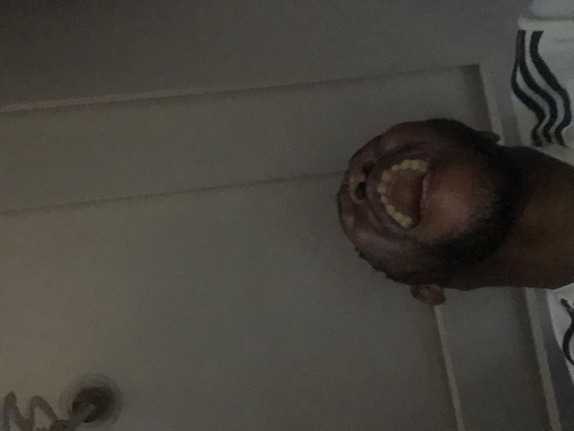 A low-angle shot looking up at a person with dark skin and a beard. The person's head is tilted back, revealing their open mouth and teeth. Part of a white shirt with black stripes is visible on the right side. The background consists of a gray wall with horizontal lines and what appears to be a doorknob in the lower left corner. The location is Unknown, Unknown.
